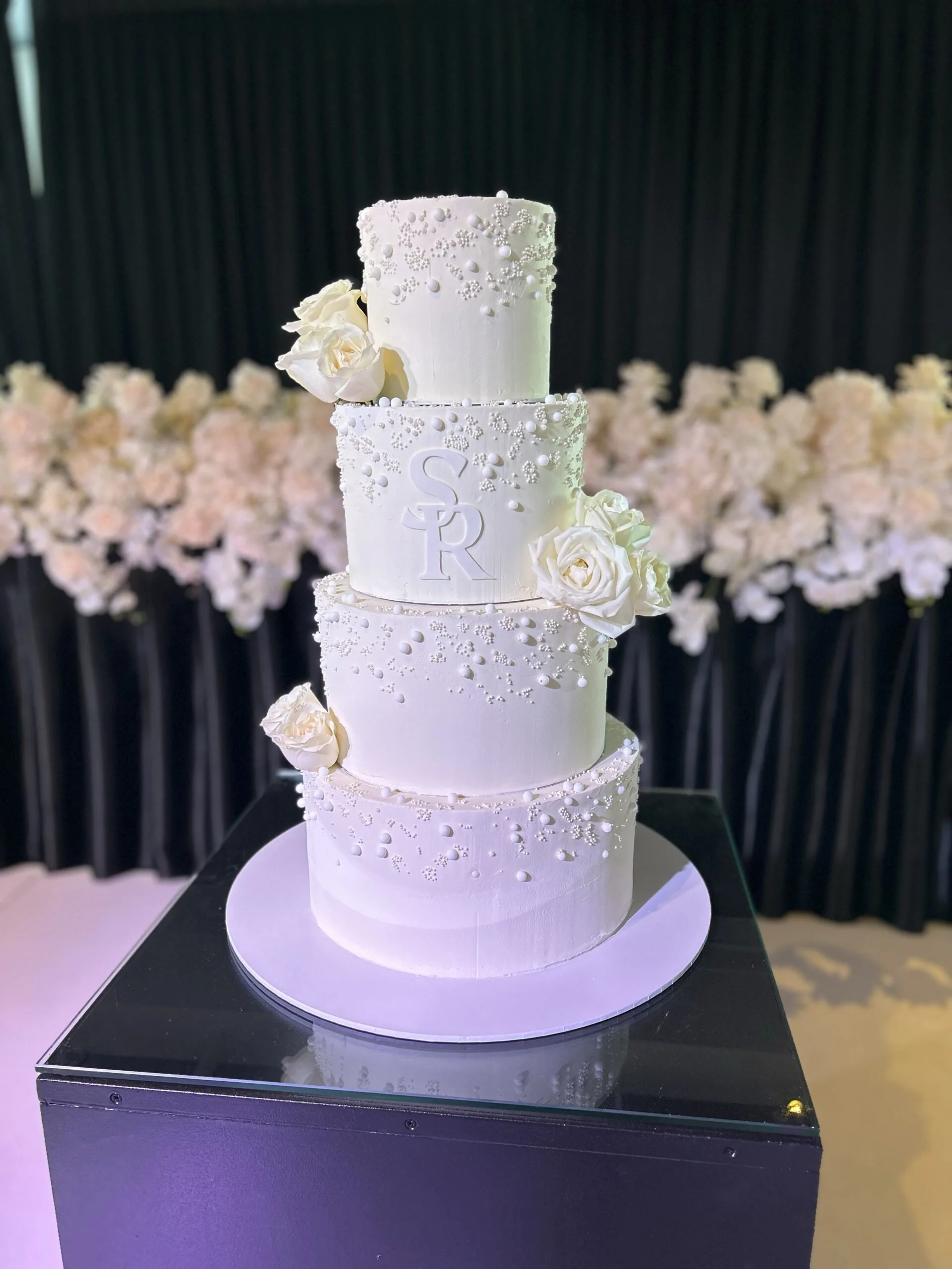 A four-tier white wedding cake decorated with white roses and pearl-like accents, with the initials S and R on the middle tier, placed on a black surface with a floral background.