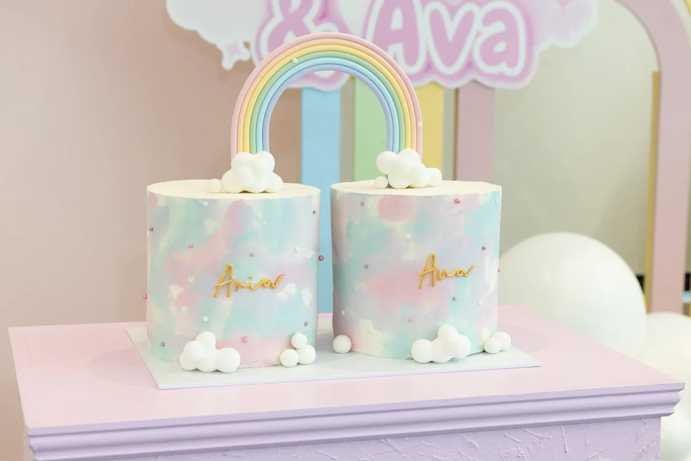 A pastel-colored cake decorated with clouds and a rainbow, with the name Aria written in gold on each side, placed on a pink table with a light pink background.