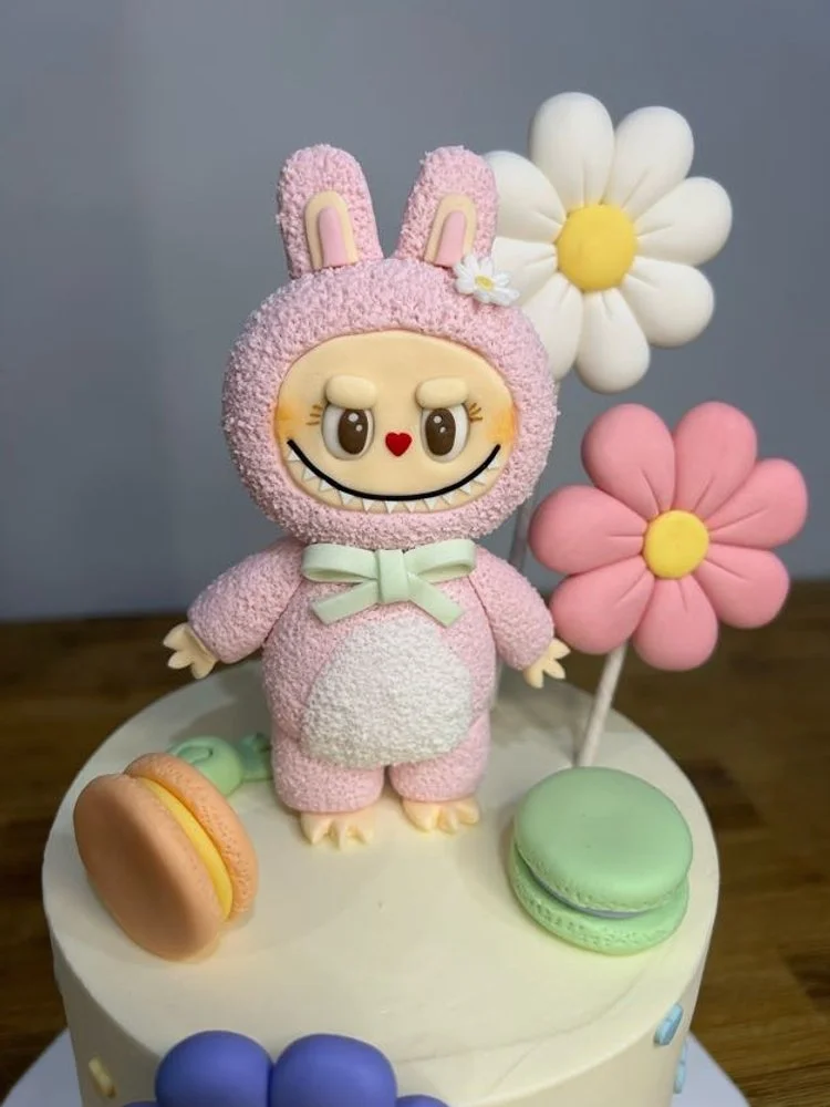 A decorated cake featuring a pink bunny character with large eyes, a red heart-shaped nose, a big smile, and a green bow. The cake is topped with fondant flowers in white and pink, and colorful macarons are placed around it.