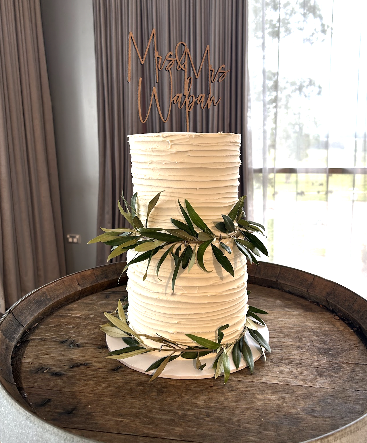 A tall wedding cake with textured white frosting and green leaves around the middle, placed on a wooden table with a sign above that reads "Mr & Mrs Wapan."
