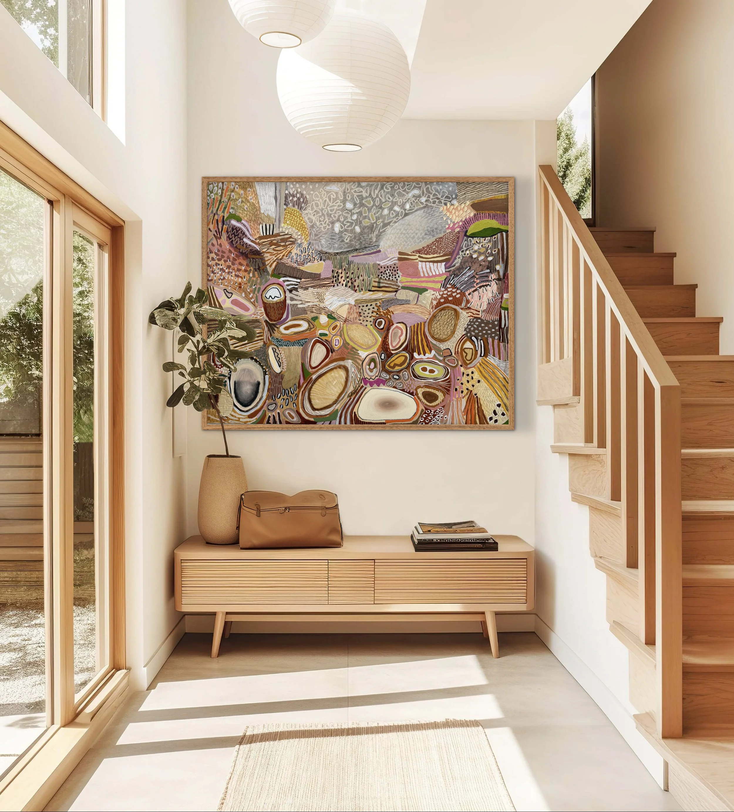 Interior of a modern home with a wooden staircase, a large colorful abstract painting on the wall, a beige console table with a vase, a purse, and some magazines, large sliding glass door, and natural light.
