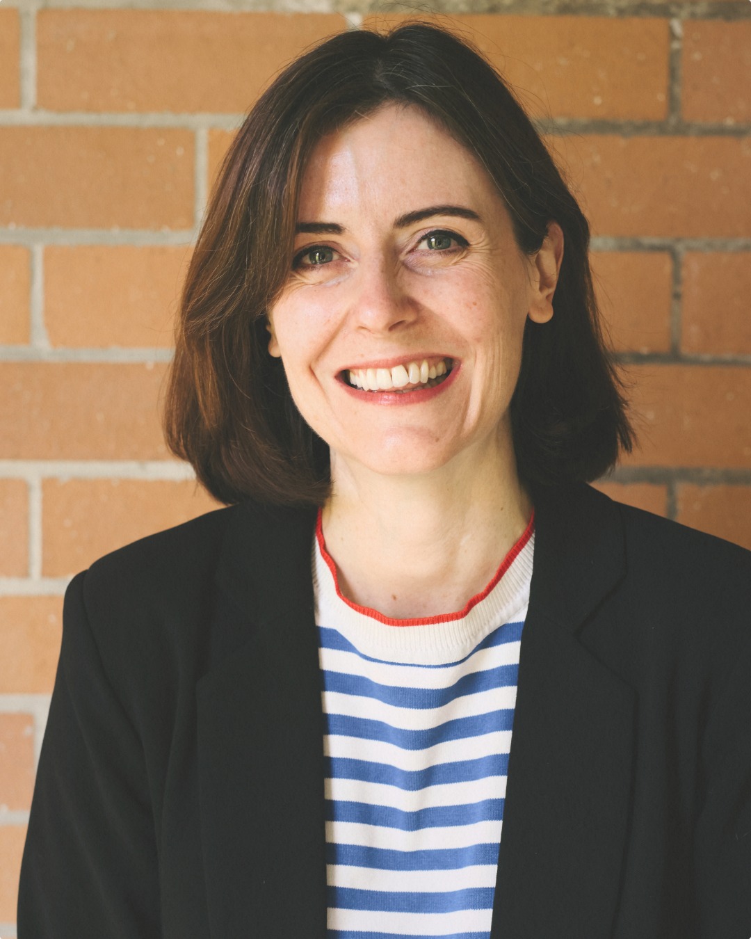 Smiling woman with dark brown hair, wearing a black blazer and a white and blue striped shirt, standing against a brick wall.
