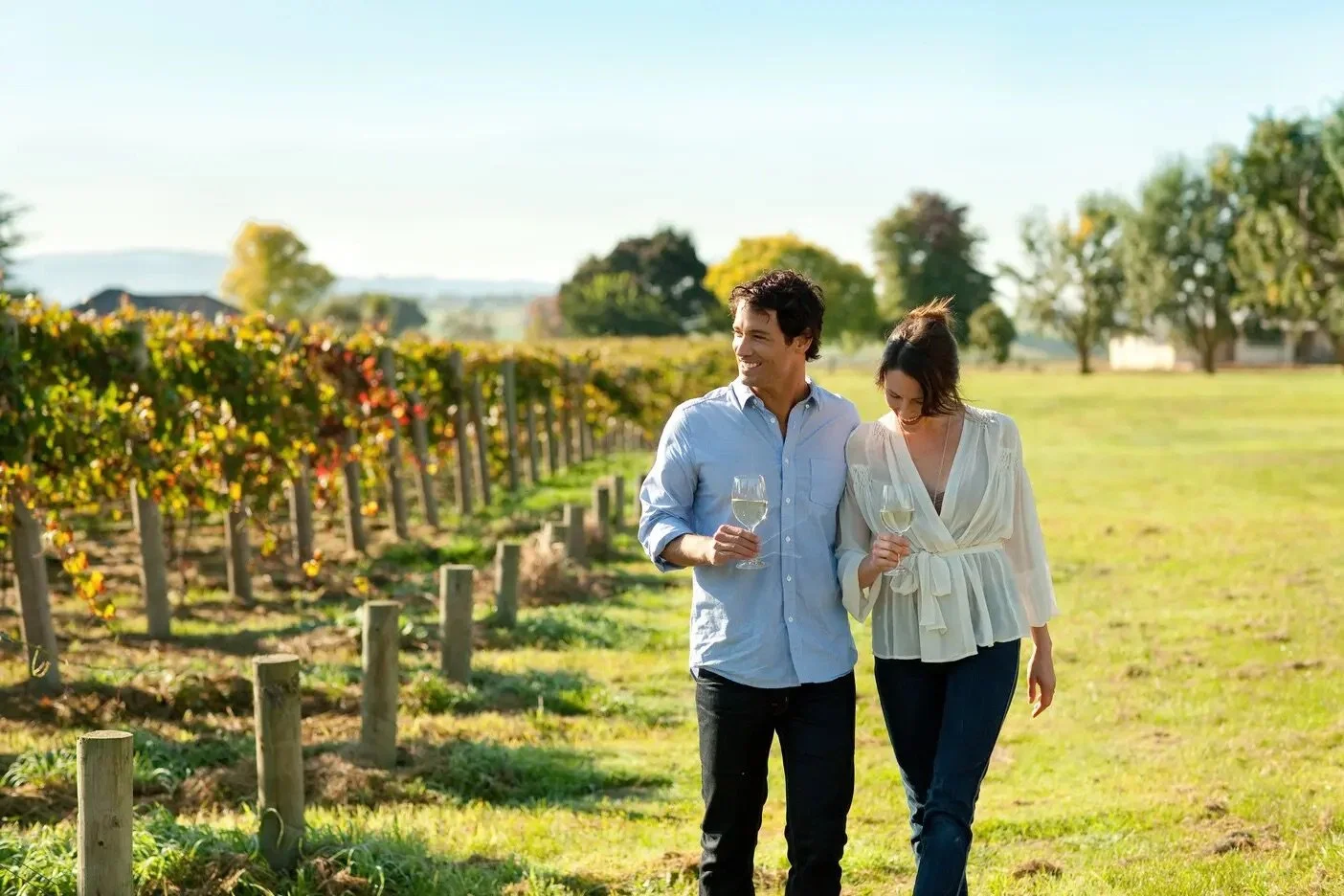 A man and woman walking through a vineyard holding glasses of white wine, smiling and enjoying the sunny day. Mornington Peninsula and Yarra Valley Wine Tours, chauffeur driven.