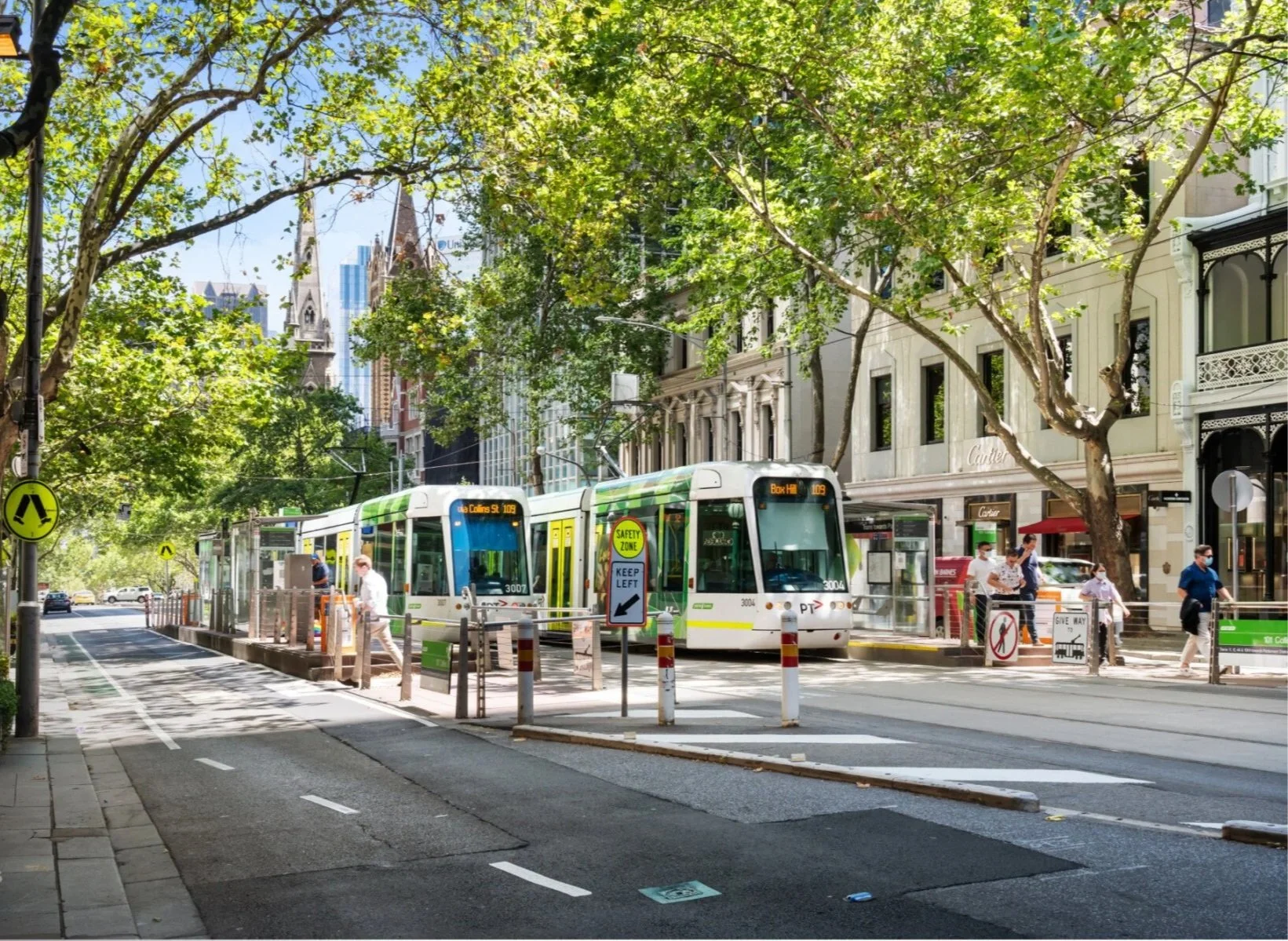 City street scene with a light rail tram stopped at a station, people walking and waiting nearby, surrounded by trees and urban buildings.