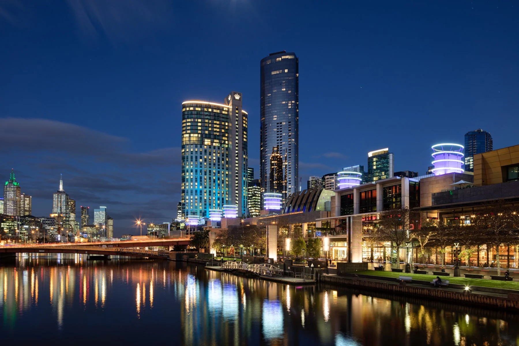 Nighttime cityscape with illuminated modern skyscrapers along a waterfront, reflecting lights on the water and a bridge in the foreground.