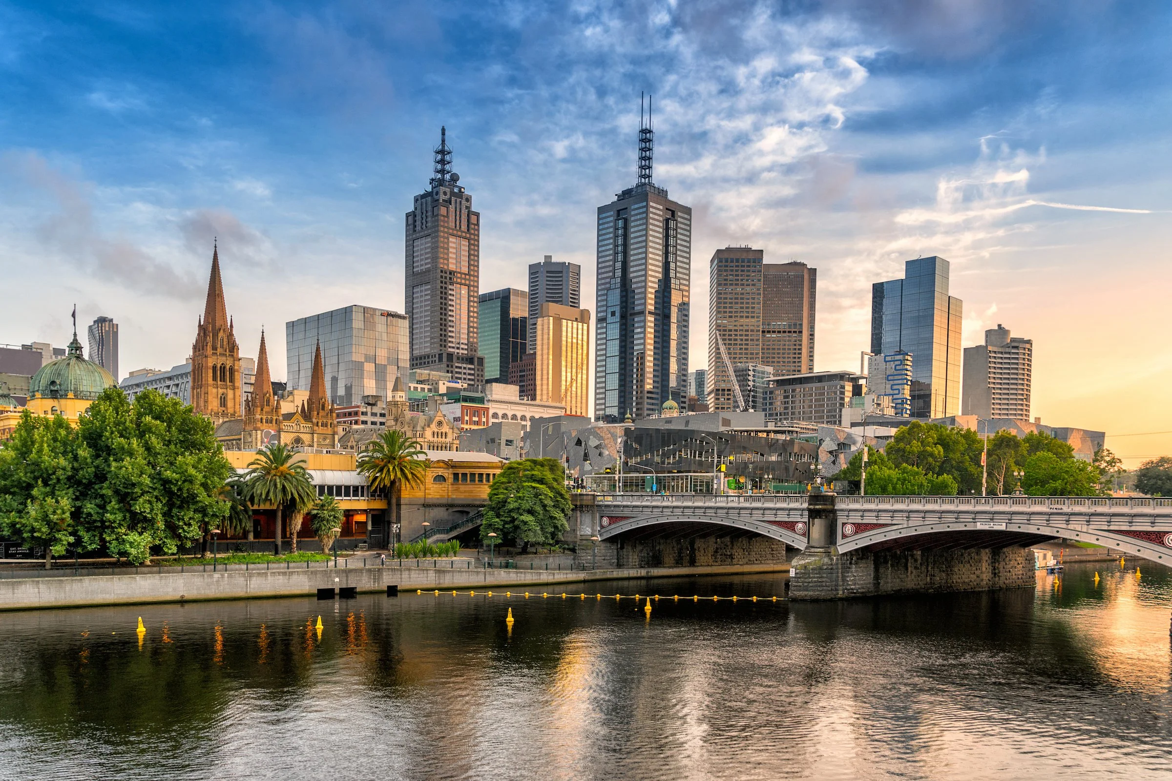 City skyline with tall skyscrapers, historic church with steeples, a bridge over the river, trees, and water reflecting the buildings in the foreground at sunset.