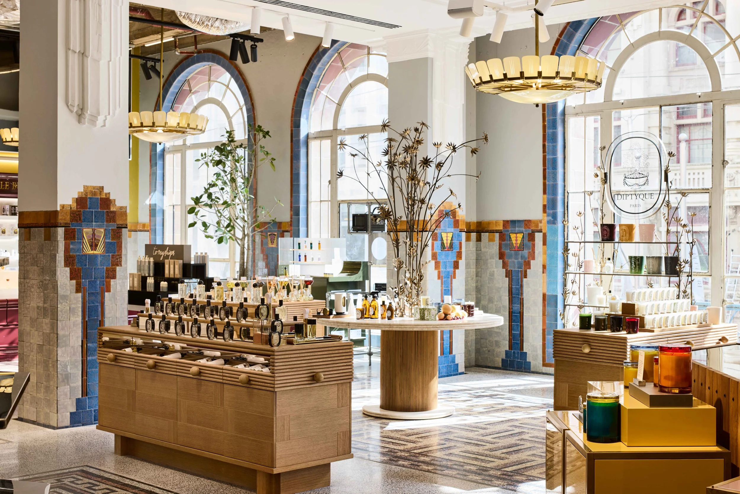 Interior view of Mecca flagship store, Bourke St Melbourne, with large arched windows, colorful tile wall accents, and modern lighting fixtures. Display shelves and tables showcase skincare and beauty products.