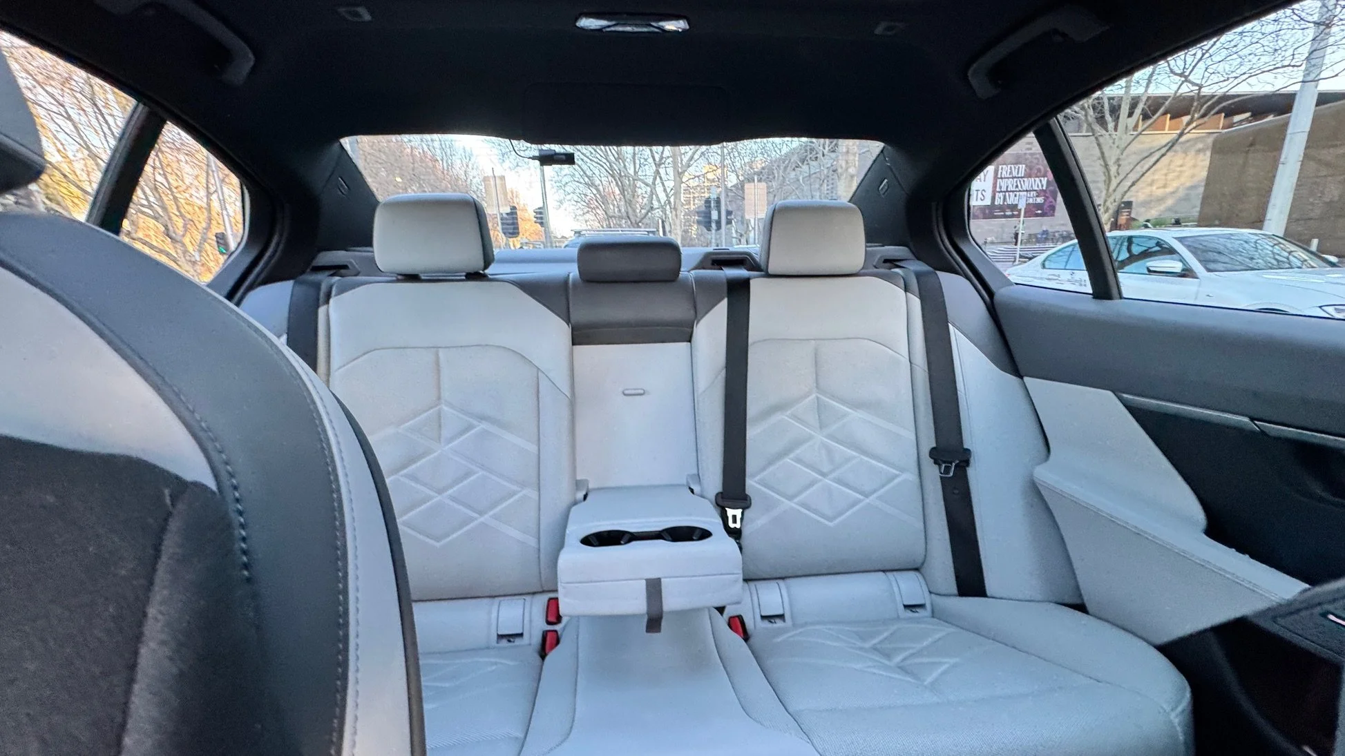 GM Drive. The interior of a modern SUV showing the back seats, with light-colored upholstery, safety seat belts, and a center armrest. Outside, there are trees and cars visible through the rear windows.