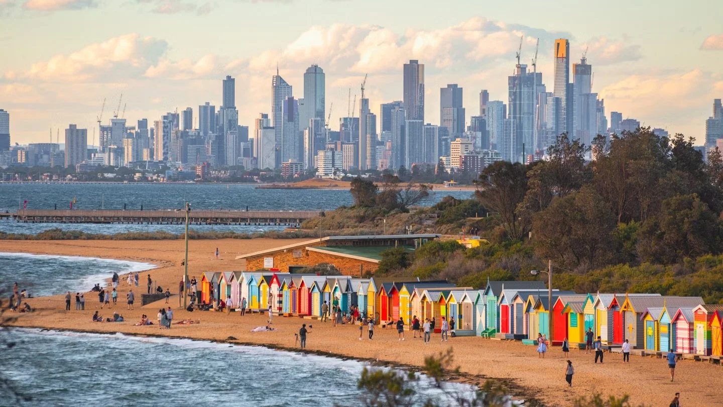 Colorful beach huts line a sandy shore with people walking and relaxing, with a city skyline featuring tall skyscrapers in the background and a body of water in the foreground. Brighton Beach boxes.