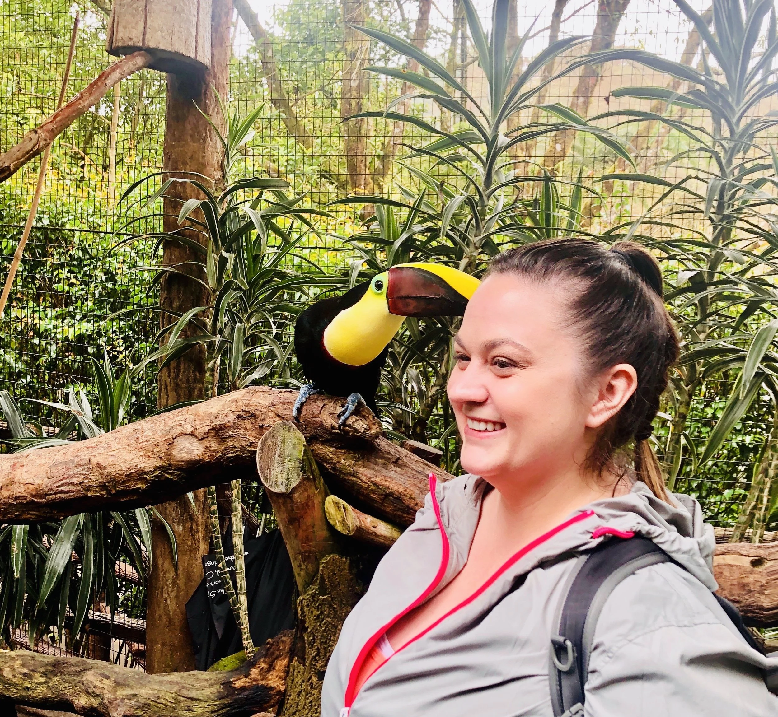 A woman smiling while standing near a toucan perched on a branch in a lush green jungle-like environment.