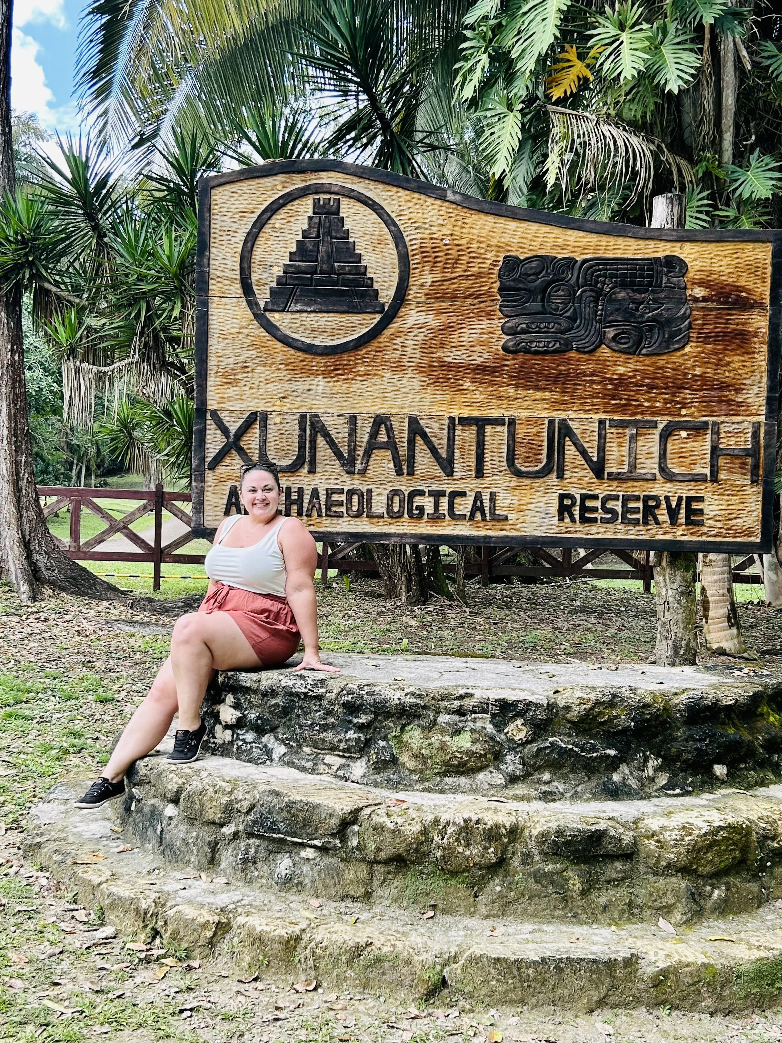 A woman sitting on stone steps next to a wooden sign that reads 'Xunantunich Archaeological Reserve', with tropical trees in the background.