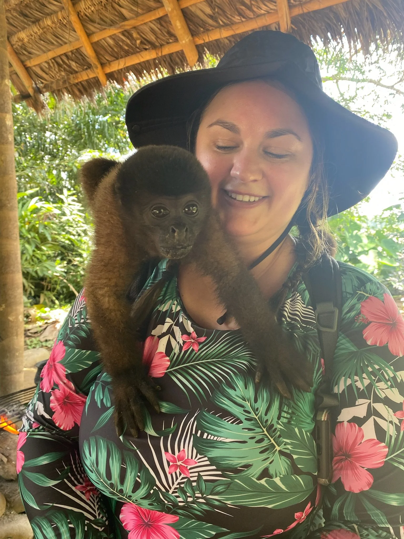 A woman in a floral tropical print shirt and black wide-brim hat smiling with a baby monkey perched on her shoulder in a lush, shaded jungle setting.
