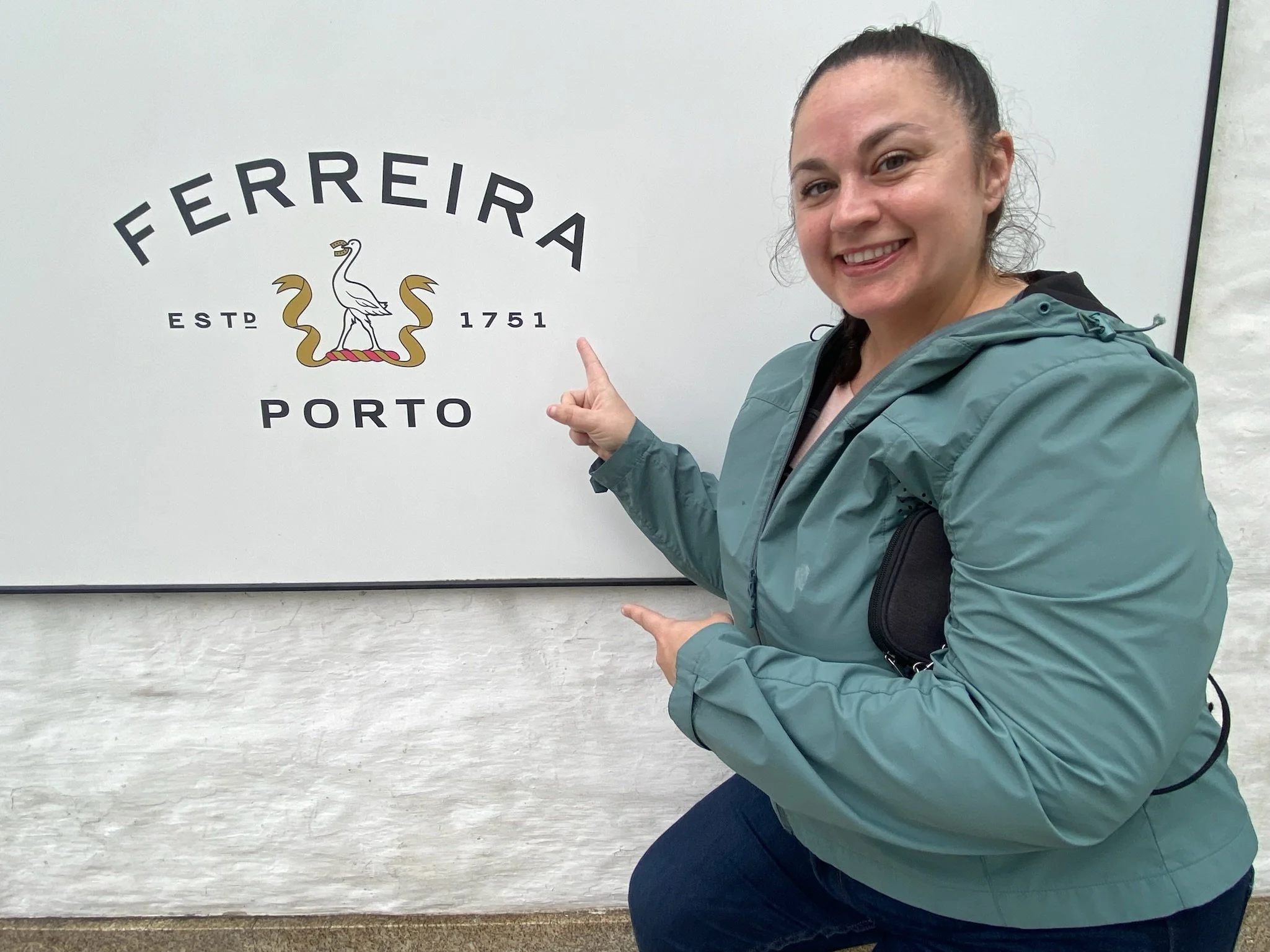 A woman in a teal jacket pointing at a sign that says "Ferreira Porto," with a logo featuring a white heron and gold decorative elements.