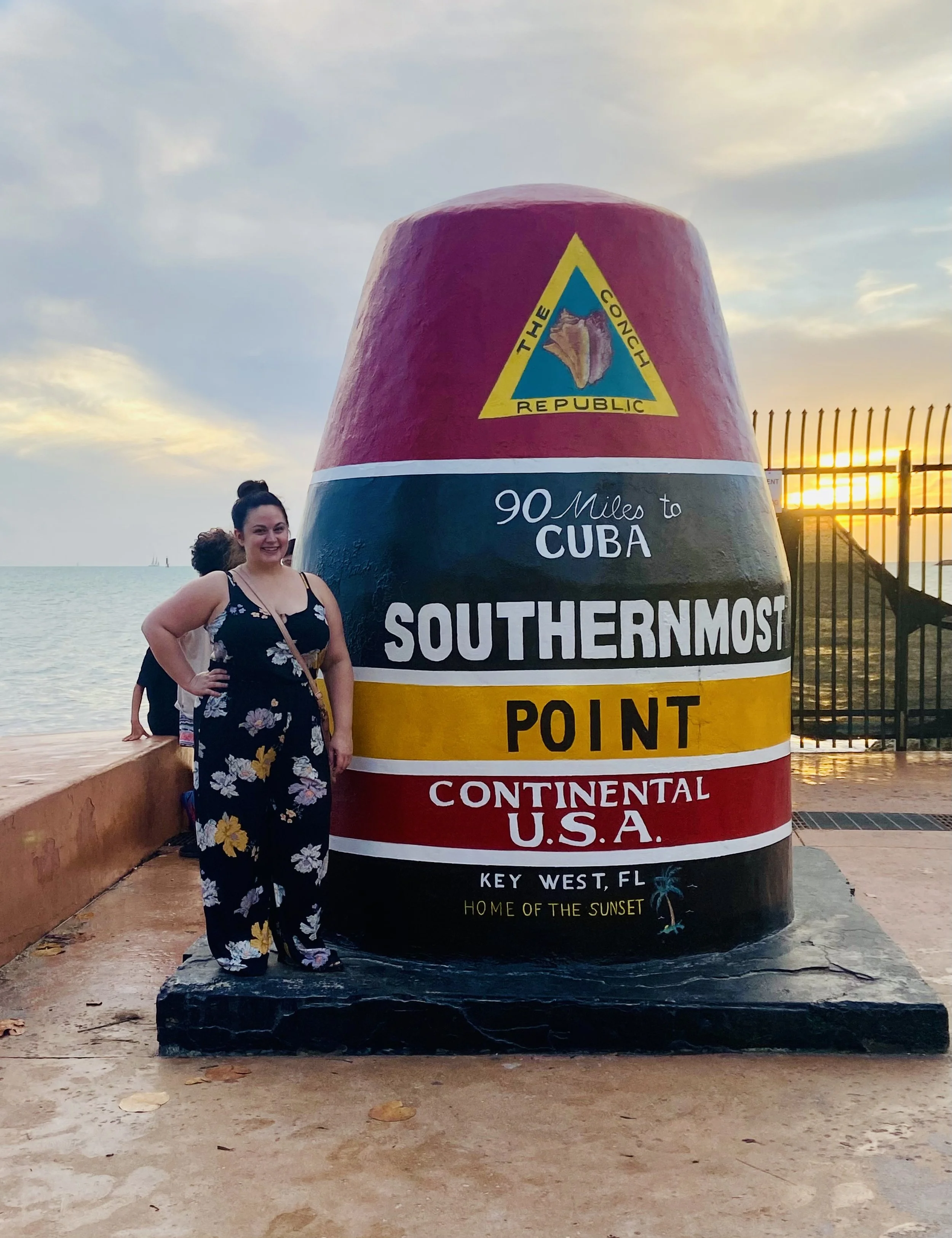 A woman standing next to a large marker monument for the southernmost point in the continental U.S., located in Key West, Florida, with the ocean and sunset in the background.