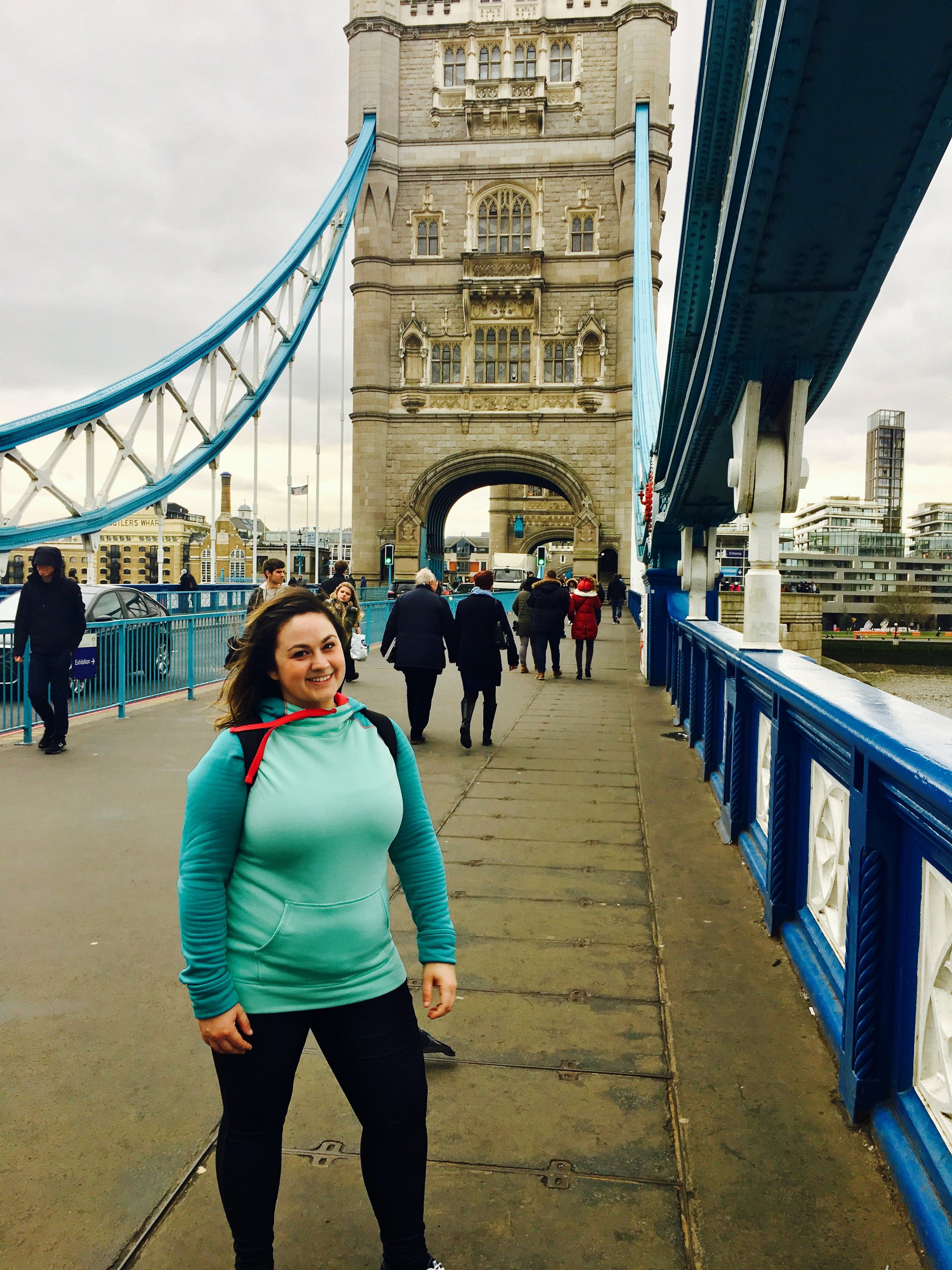 Smiling woman in teal hoodie and black pants standing on Tower Bridge in London, England, with tourists walking in the background.