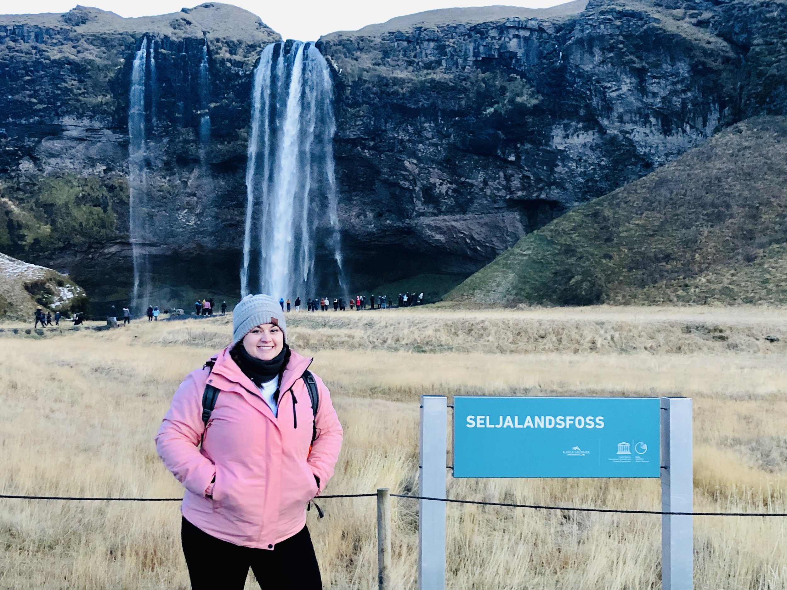 Smiling woman in pink jacket and gray hat standing in front of the Seljalandsfoss waterfall in Iceland.