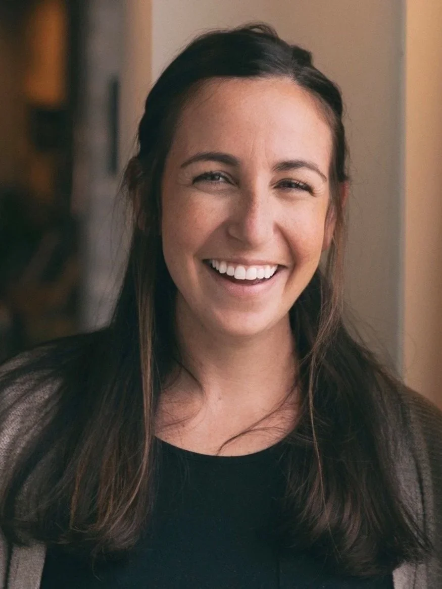 A woman with long dark hair smiling, wearing a black top and a grey cardigan, in an indoor setting.
