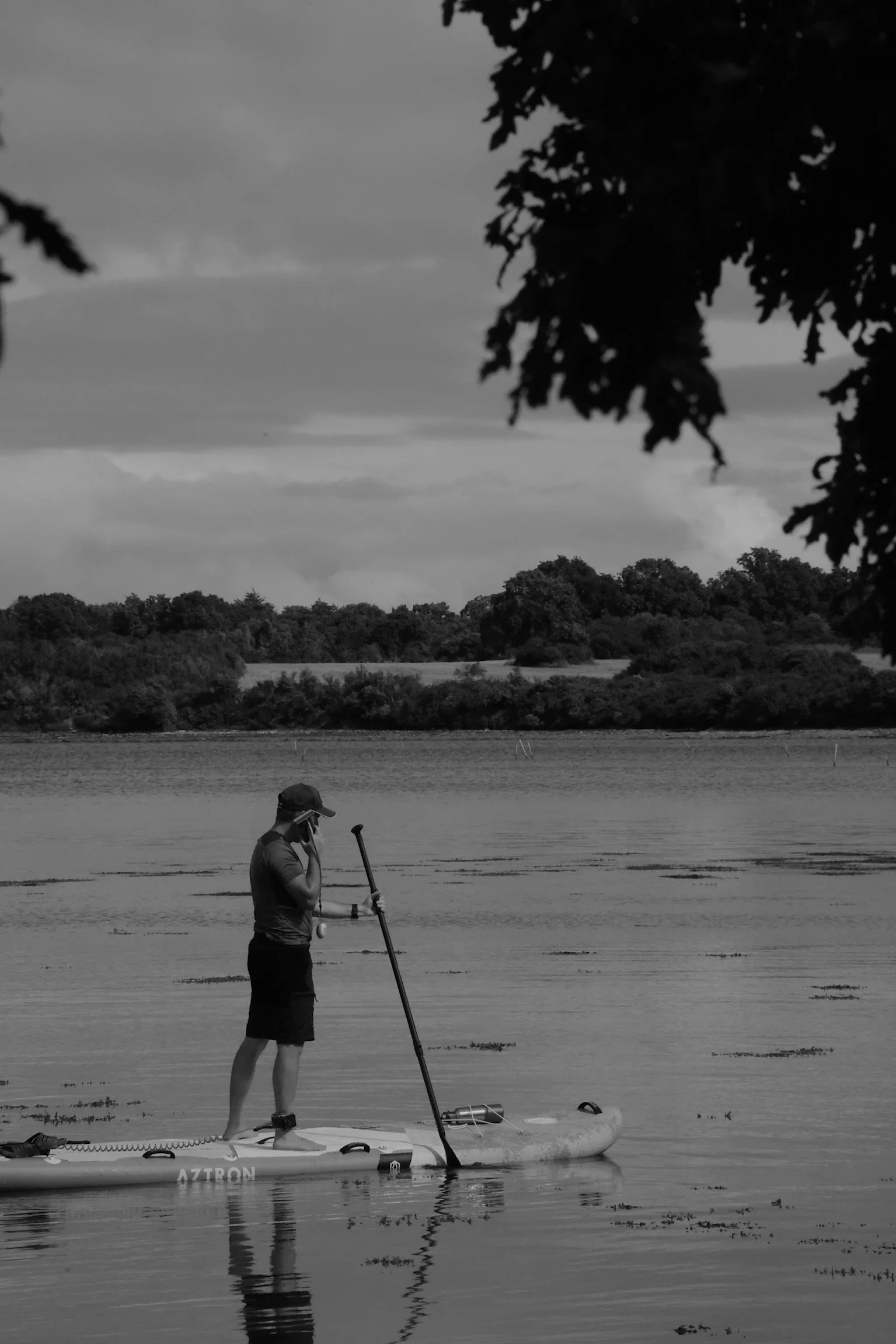 Person on paddleboard in a lake, wearing a cap, talking on a phone with trees in the background.
