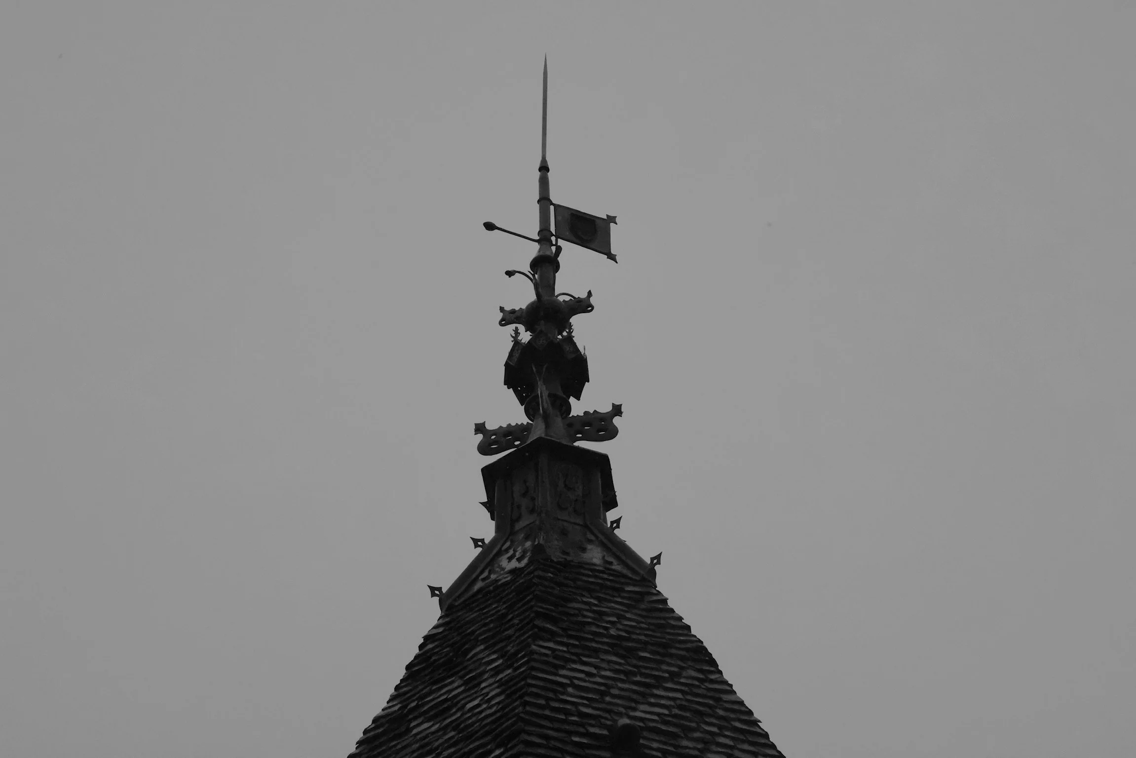 A black and white photo of a pointed tower roof with an ornate metal weather vane at the top.