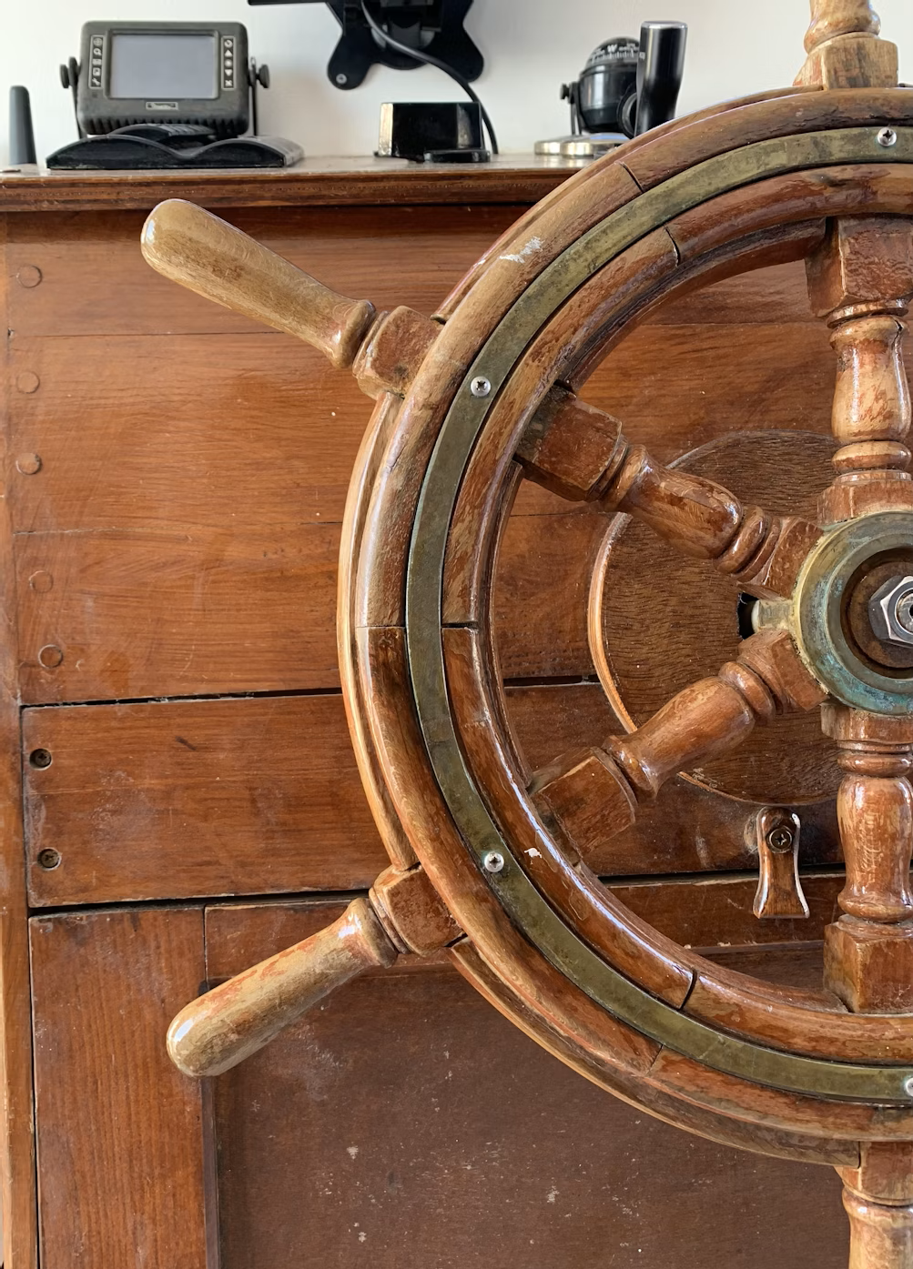 Close-up of a wooden ship steering wheel on a wooden surface with electronic equipment in the background.