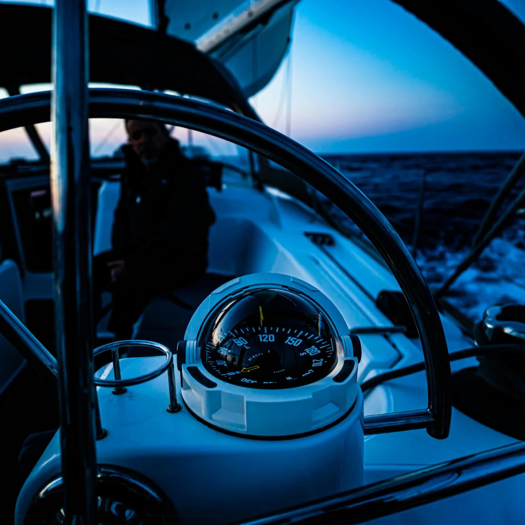 Close-up of a boat's navigational instrument display showing a compass, with a blurred person sitting in the background and ocean surroundings in the evening or early morning light.