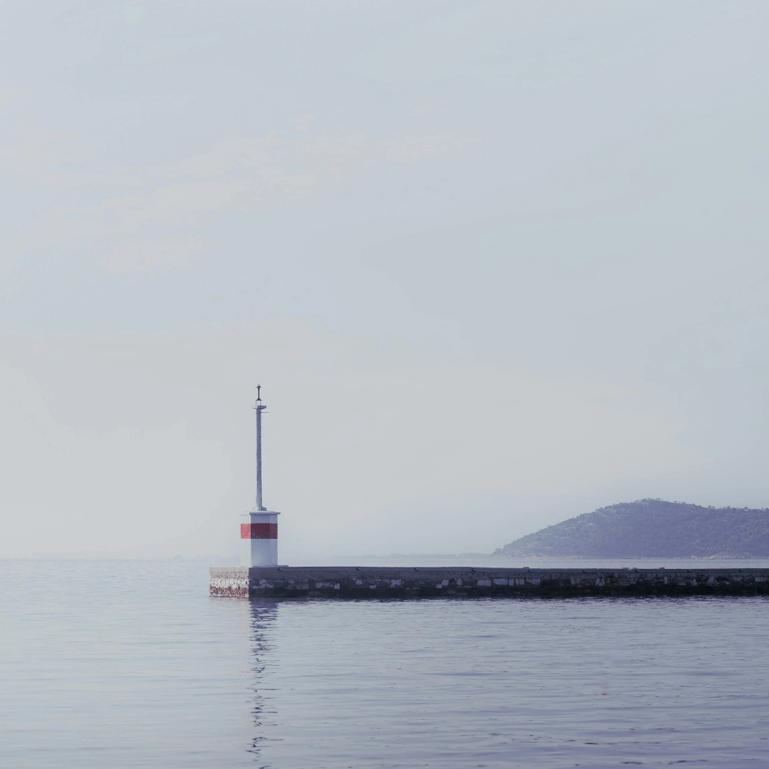 A lighthouse on a pier extending into calm water with a distant landform in the background.