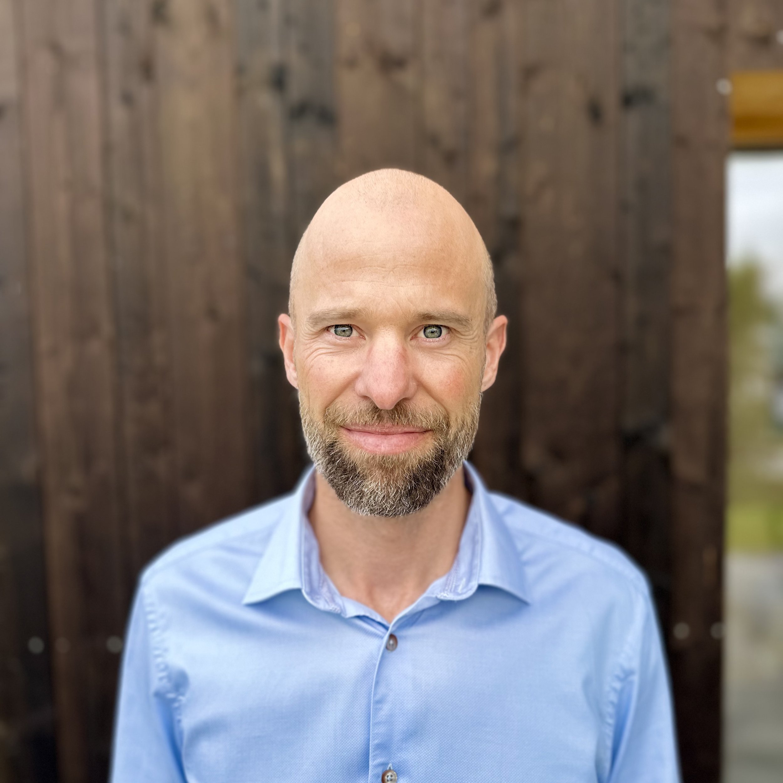A middle-aged man with a beard and blue eyes wearing a light blue button-up shirt, standing outdoors in front of a wooden wall.