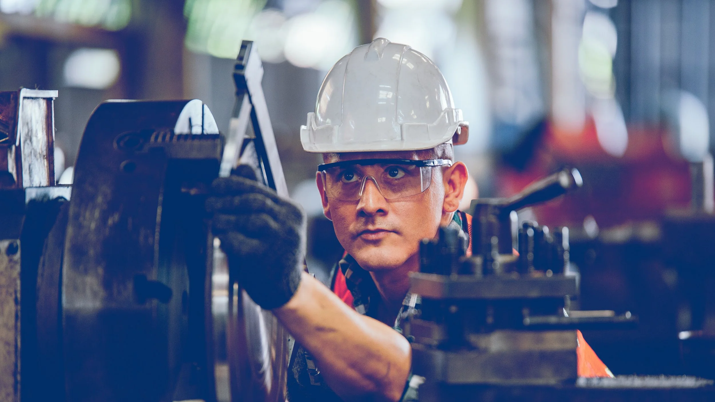 A male industrial worker wearing safety glasses, a white helmet, and a safety vest is working on a machine in a factory or workshop environment.