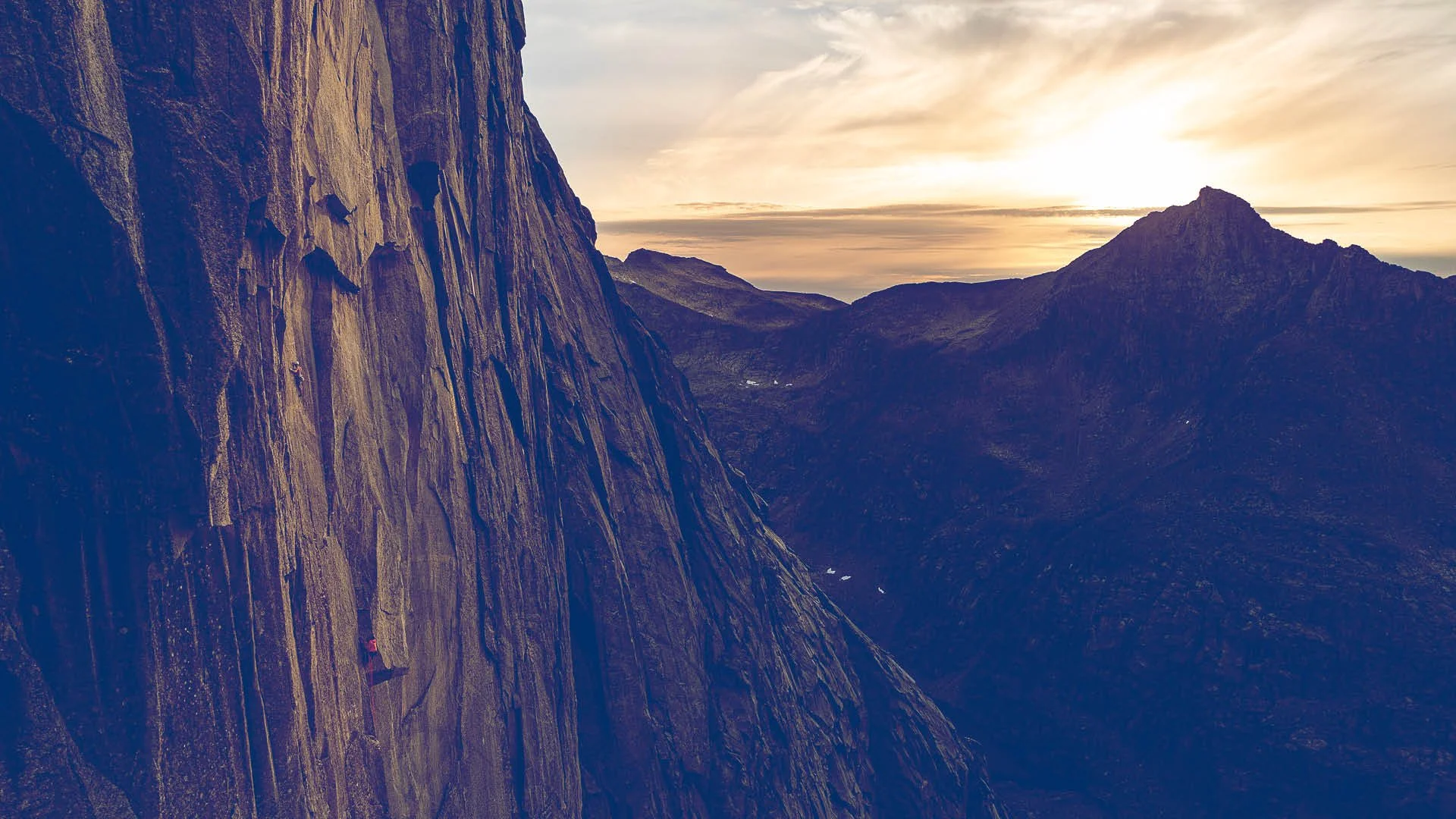 A climber ascending a tall, steep rock face during sunset with mountain peaks in the distance.
