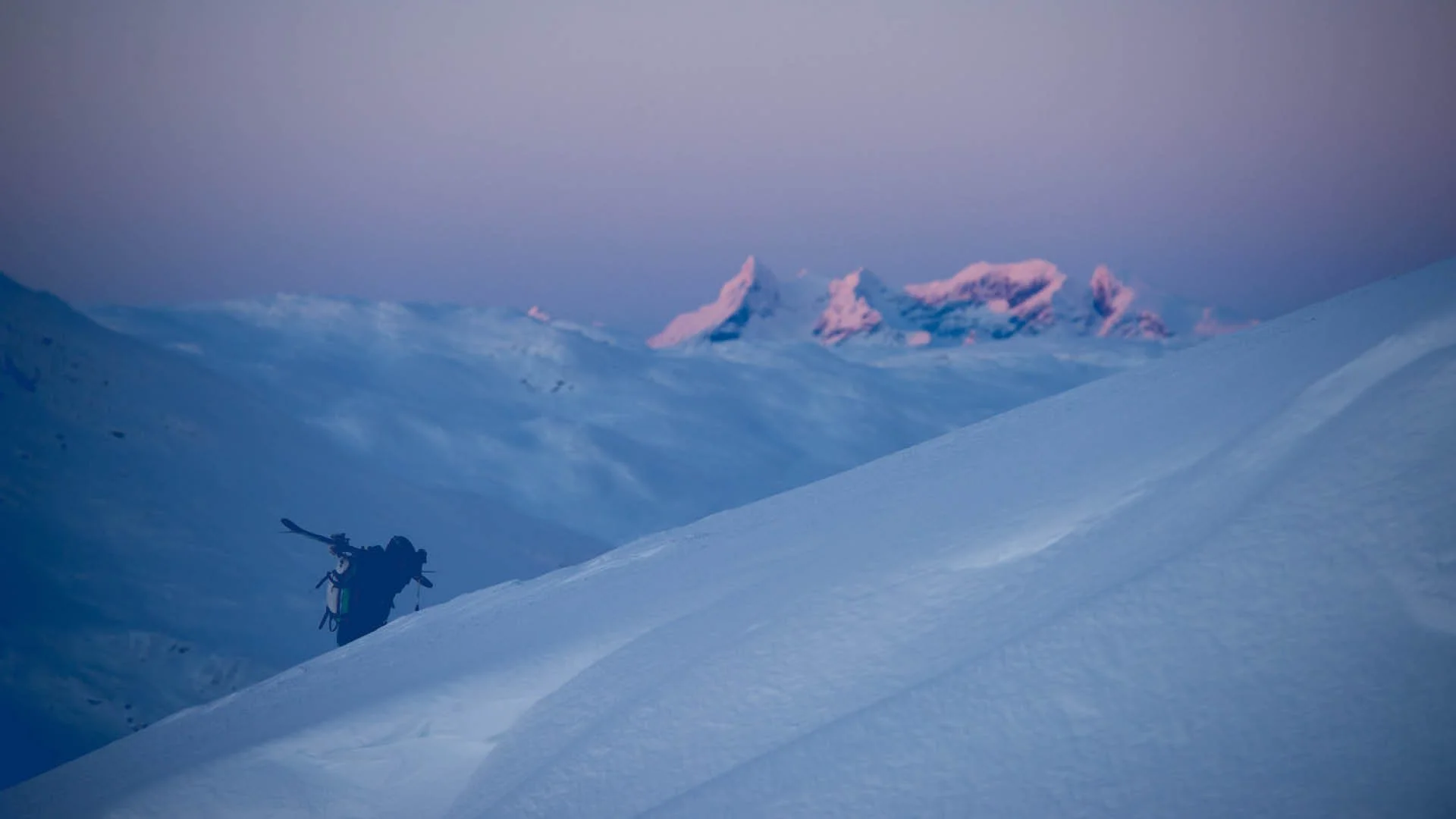 A lone skier with ski gear walking up a snowy mountain slope during dusk, with snow-covered peaks in the background under a pinkish-purple sky.
