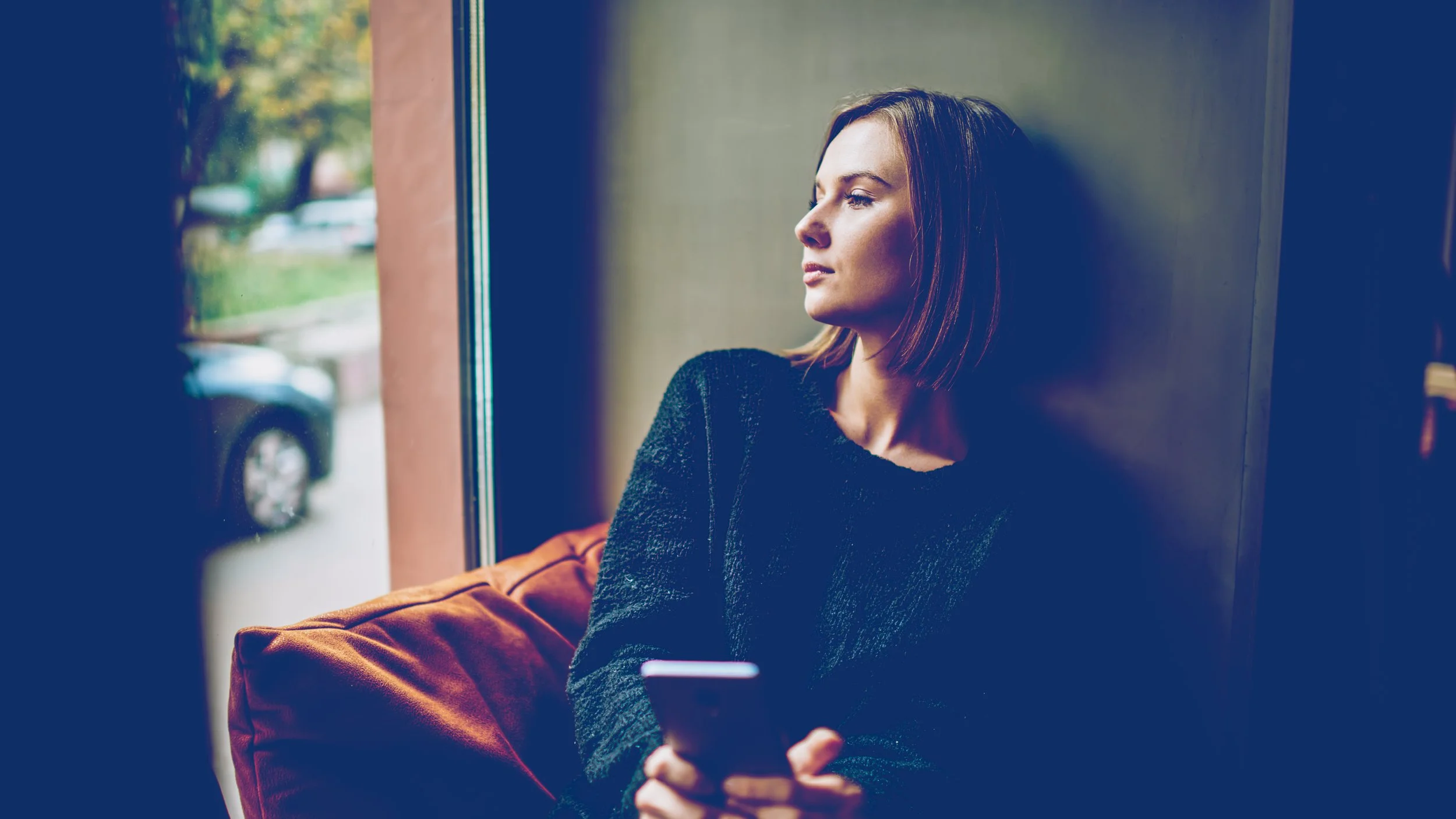 Woman with brown hair looking out the window, holding a smartphone, sitting on an orange cushion.