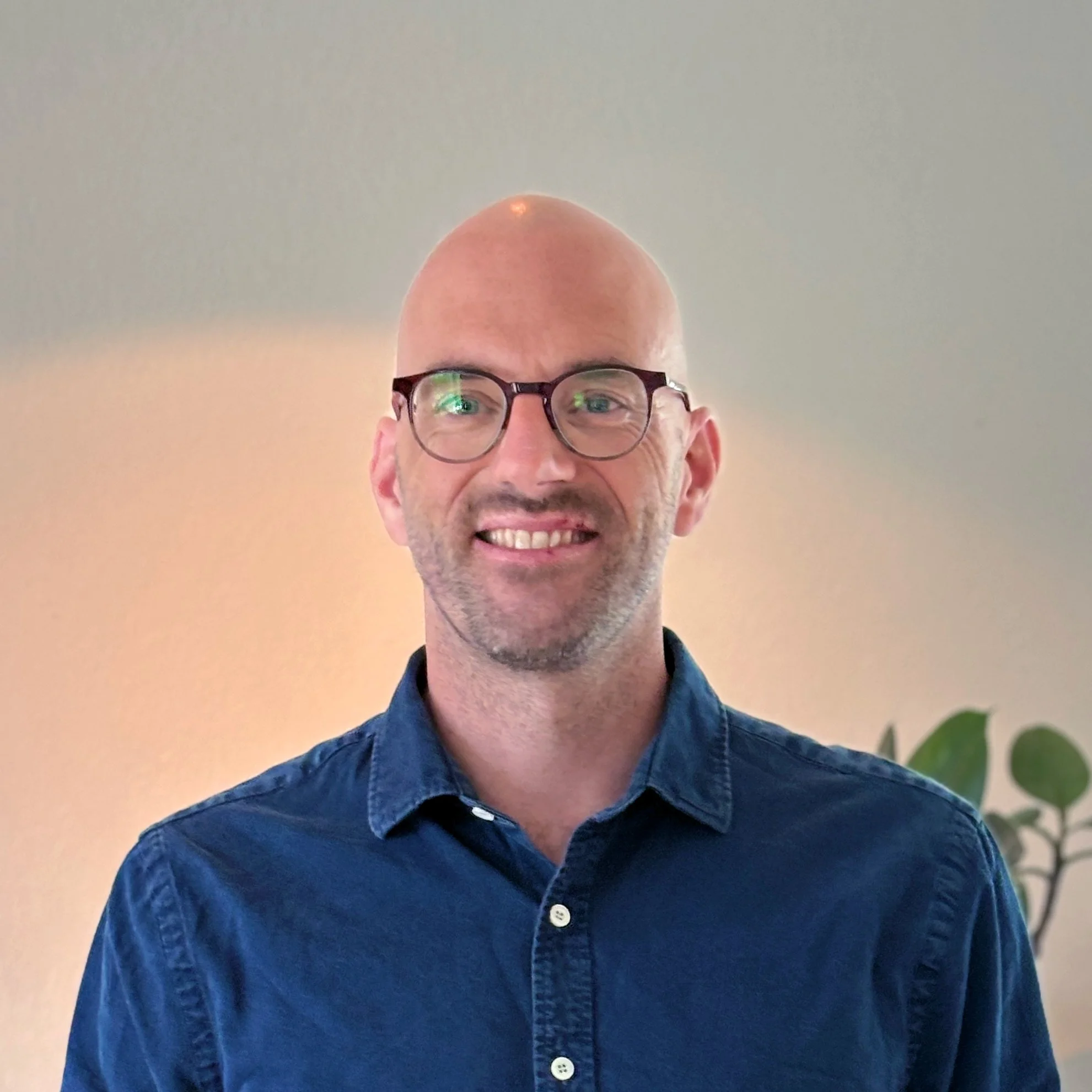 Portrait of a smiling man with glasses wearing a blue button-up shirt, standing indoors near a plant against a neutral background.