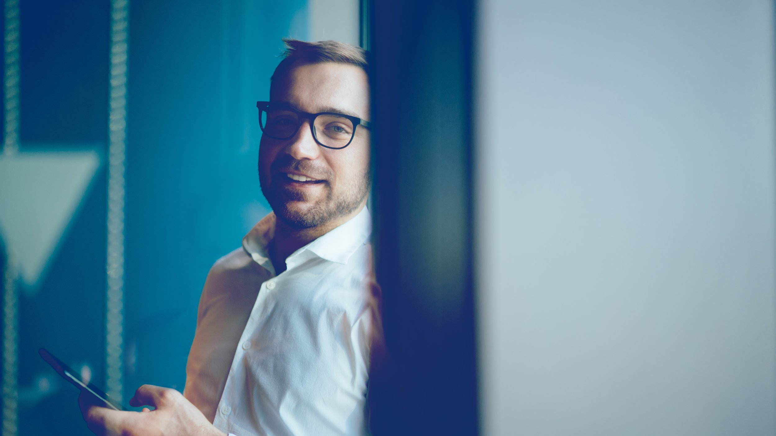 A man with glasses and a white shirt smiling while looking at the camera inside an office environment.