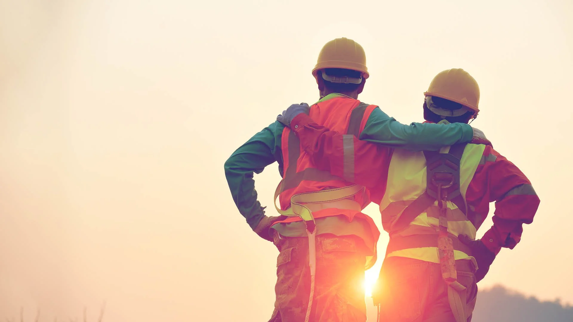 Two construction workers in safety gear standing together outdoors at sunset, with one having an arm around the other's shoulder.