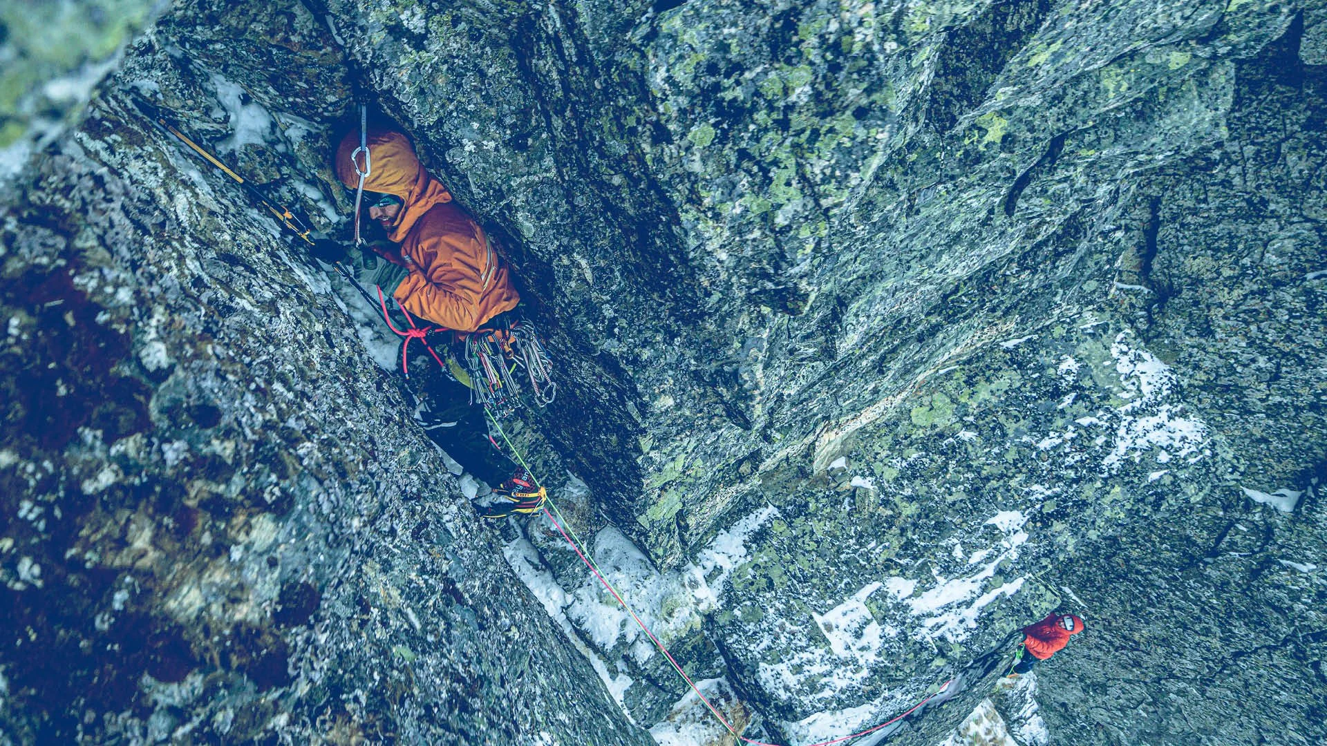 Two climbers ascending a steep, rocky mountain face with snow patches, secured with safety ropes and gear.