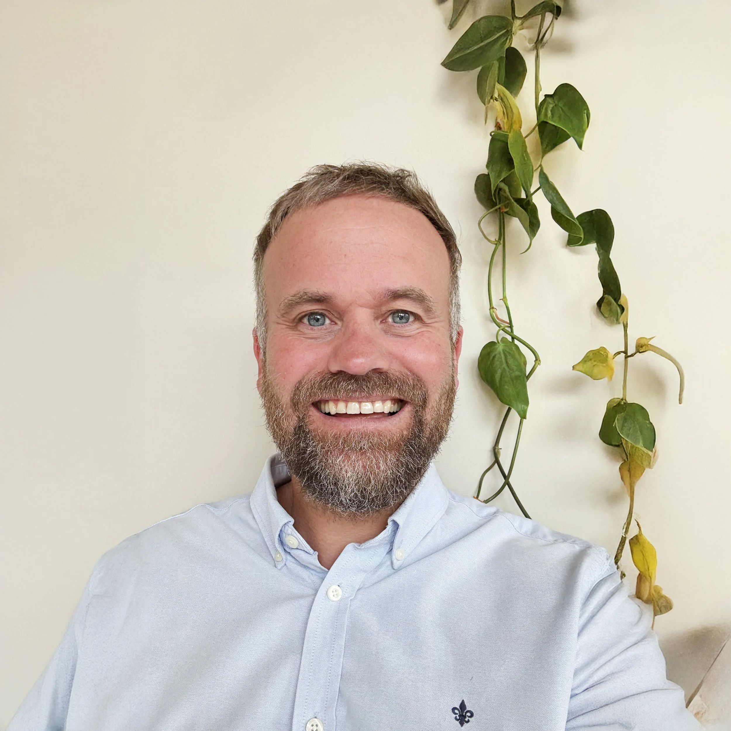 A smiling man with a beard and blue eyes wearing a light blue button-up shirt, standing in front of a cream-colored wall with green trailing plants.