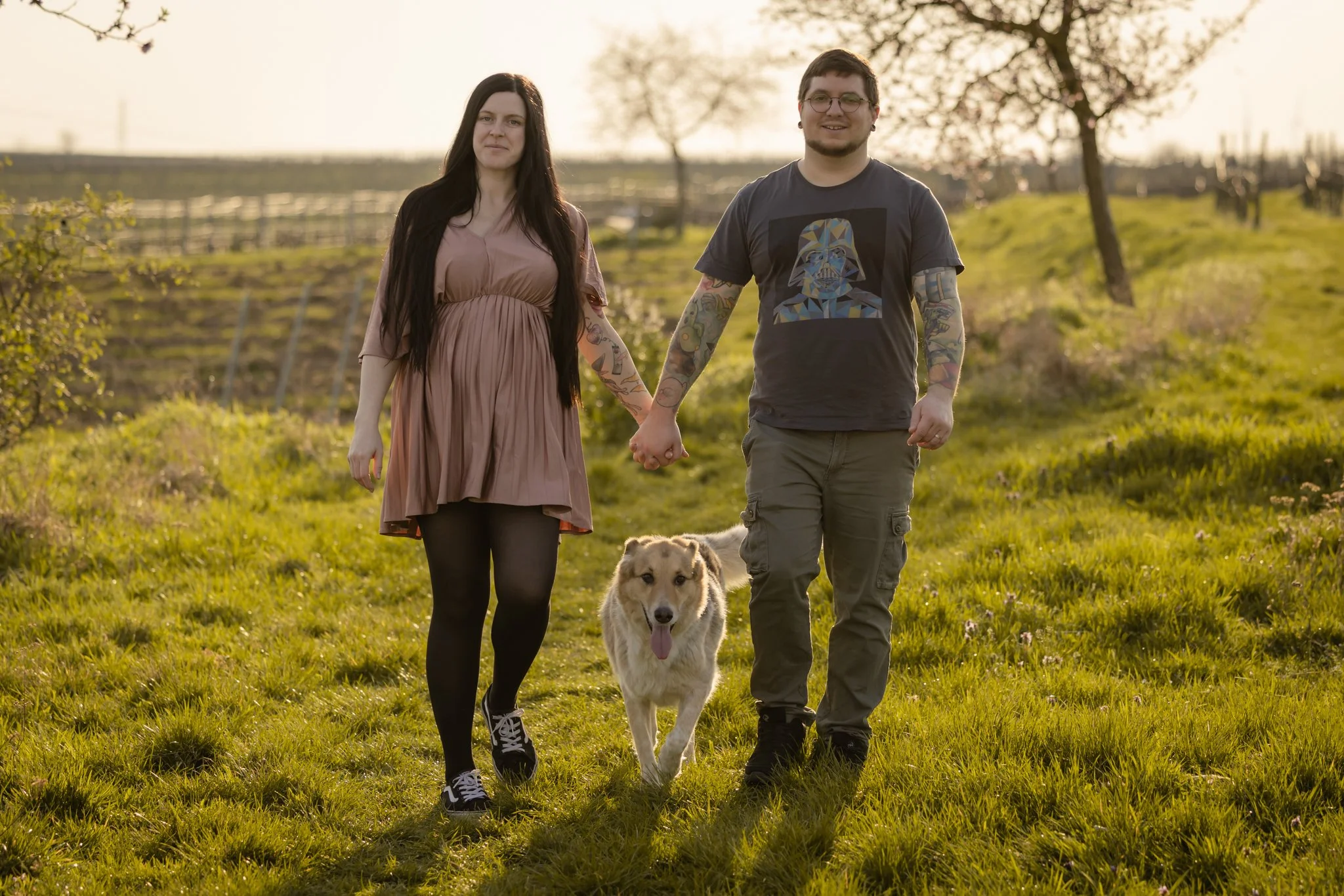 Ein Mann und eine Frau laufen an einem sonnigen Tag im Grünen, Hand in Hand, mit einem Hund. Im Hintergrund sind Bäume und eine ländliche Landschaft sichtbar.
