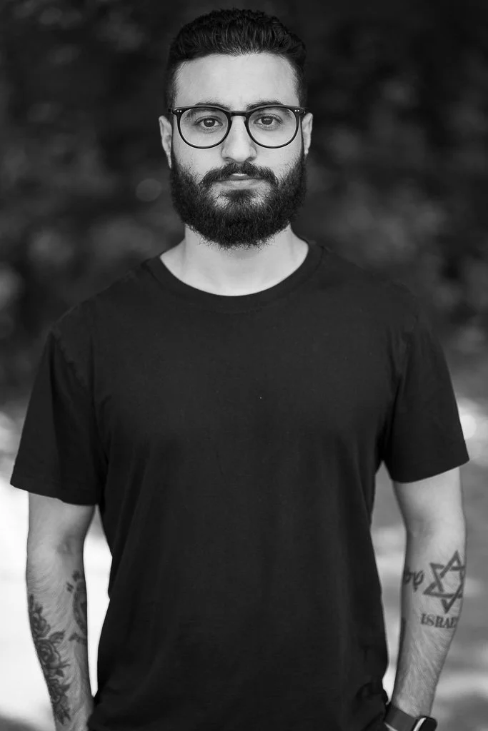 Junger Mann mit Brille, dunklem Haar, Vollbart und Tattoos, trägt schwarzen T-Shirt, blickt in die Kamera, unscharfer Hintergund