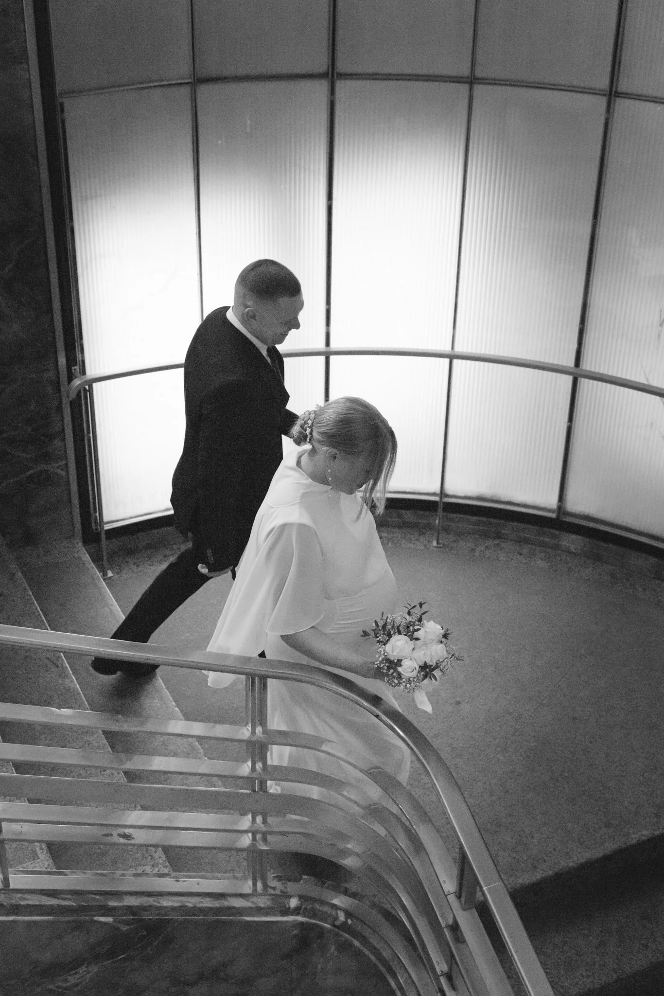 Ein Hochzeitsfoto eines Paares, das die Treppe im Gebäude hinuntergeht. Die Braut trägt ein weißes Hochzeitskleid und hält einen Blumenstrauß, der Bräutigam trägt einen schwarzen Anzug.