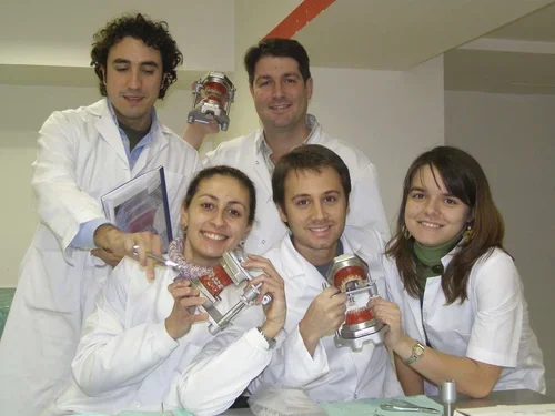 Cinco personas en batas blancas, sonriendo y sosteniendo objetos de laboratorio en un entorno interior.
