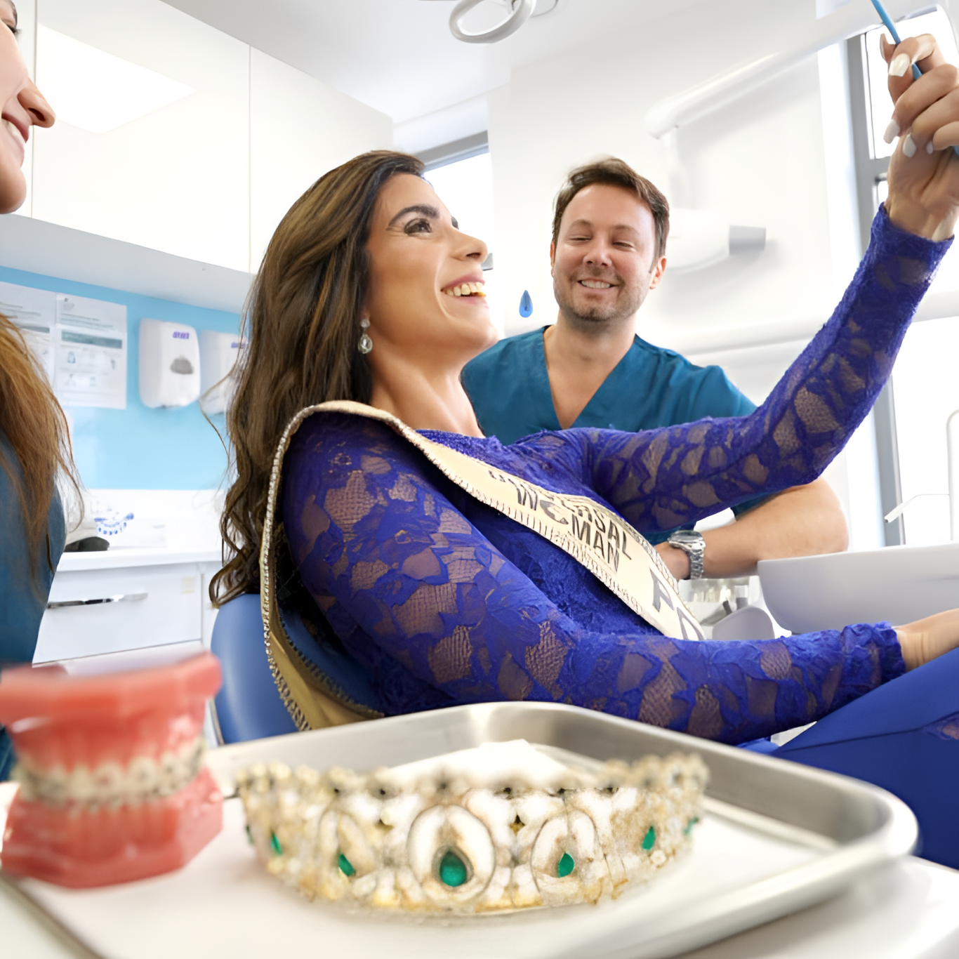 Una mujer en un consultorio dental con un vestido de encaje azul y una banda que dice 'Reina', sonriendo mientras el dentista y otra persona la acompañan. En primer plano, un modelo de mandíbula y una corona dental en una bandeja.