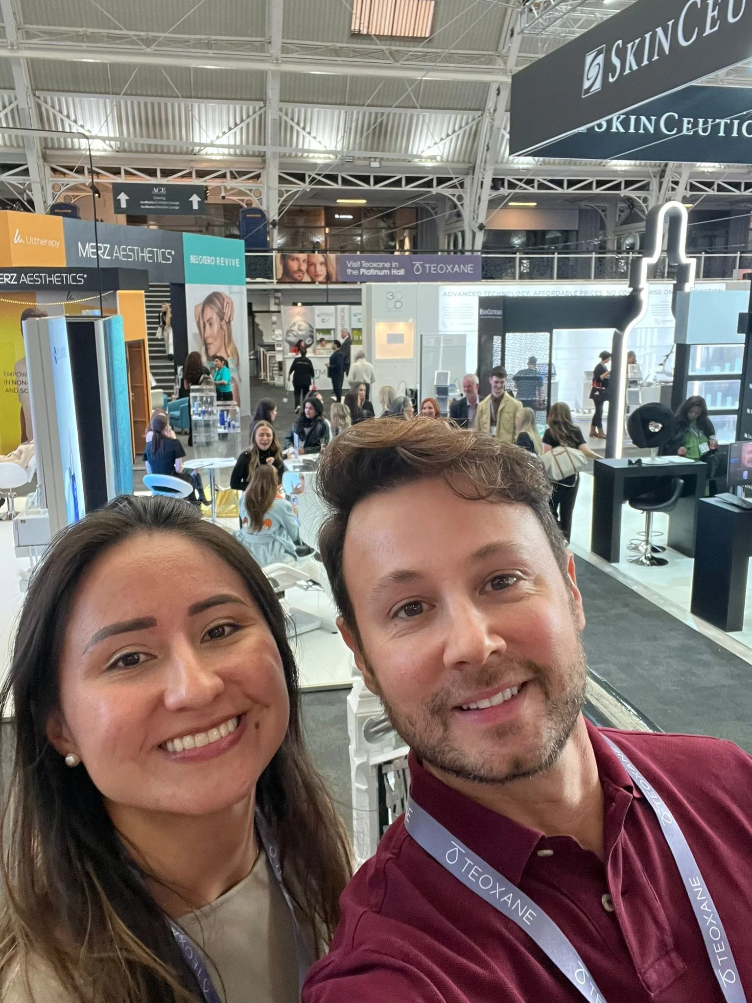 Dos personas sonrientes tomando una selfie en una exposición de estética y tecnología en un recinto moderno y concurrido.