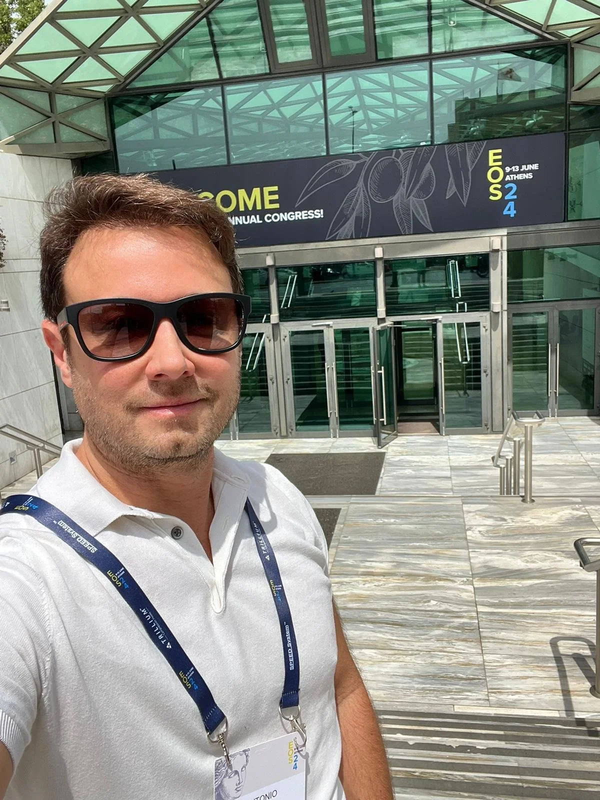 Hombre con gafas de sol y camiseta blanca tomadas en la entrada de un edificio con un cartel que dice 'WELCOME' y detalles del congreso en Atenas, Grecia.