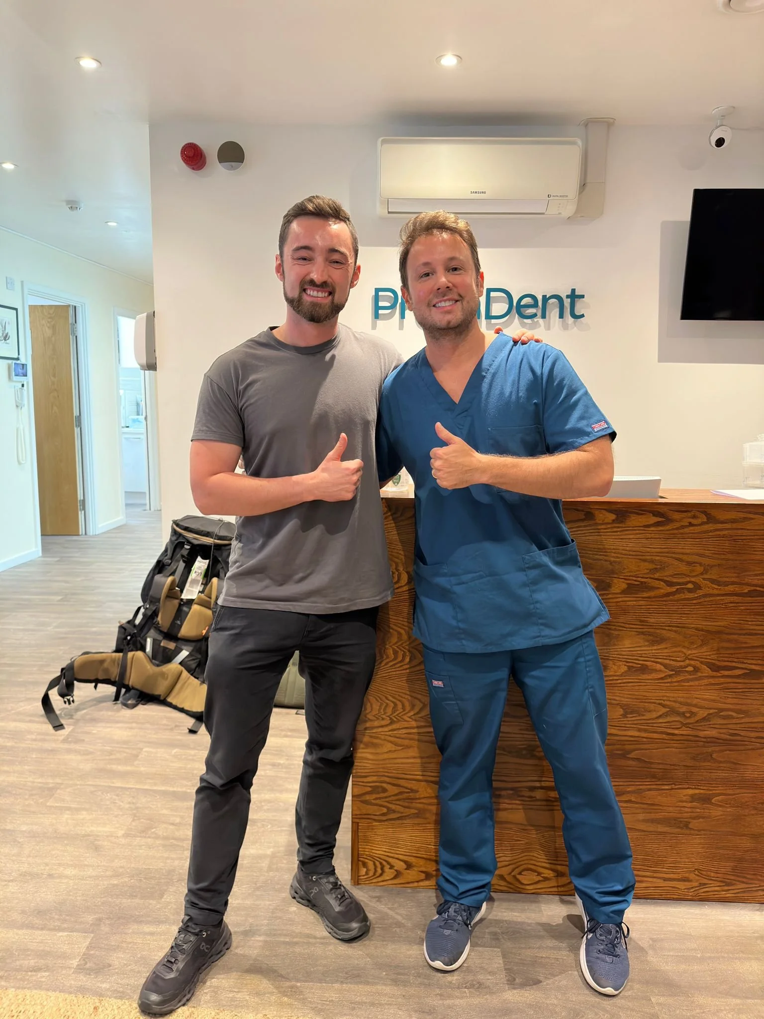 Dos hombres, uno vestido con ropa casual y el otro con uniforme de dentista, posando sonriendo y dando pulgares en la recepción de una clínica dental.