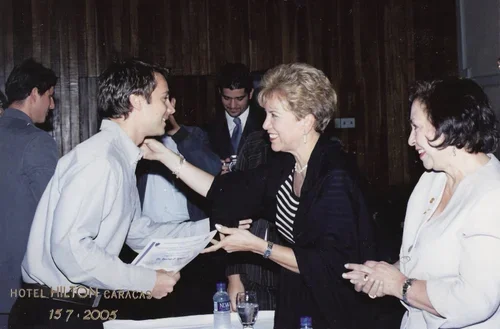 Un grupo de personas en una reunión, sonriendo y saludándose, en una sala con paredes de madera, en una ocasión formal o de negocios.