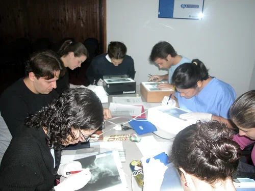 Personas en una mesa realizando tareas, probablemente en un entorno de estudio o trabajo, con materiales como libros, papeles y equipo tecnológico.