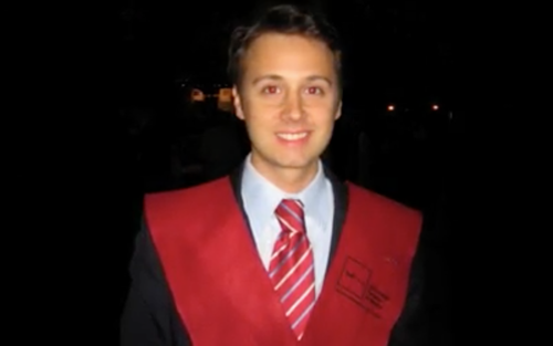 Joven sonriendo en una ceremonia de graduación con toga y birrete negra, llevando una camisa blanca y corbata a rayas rojas y blancas.