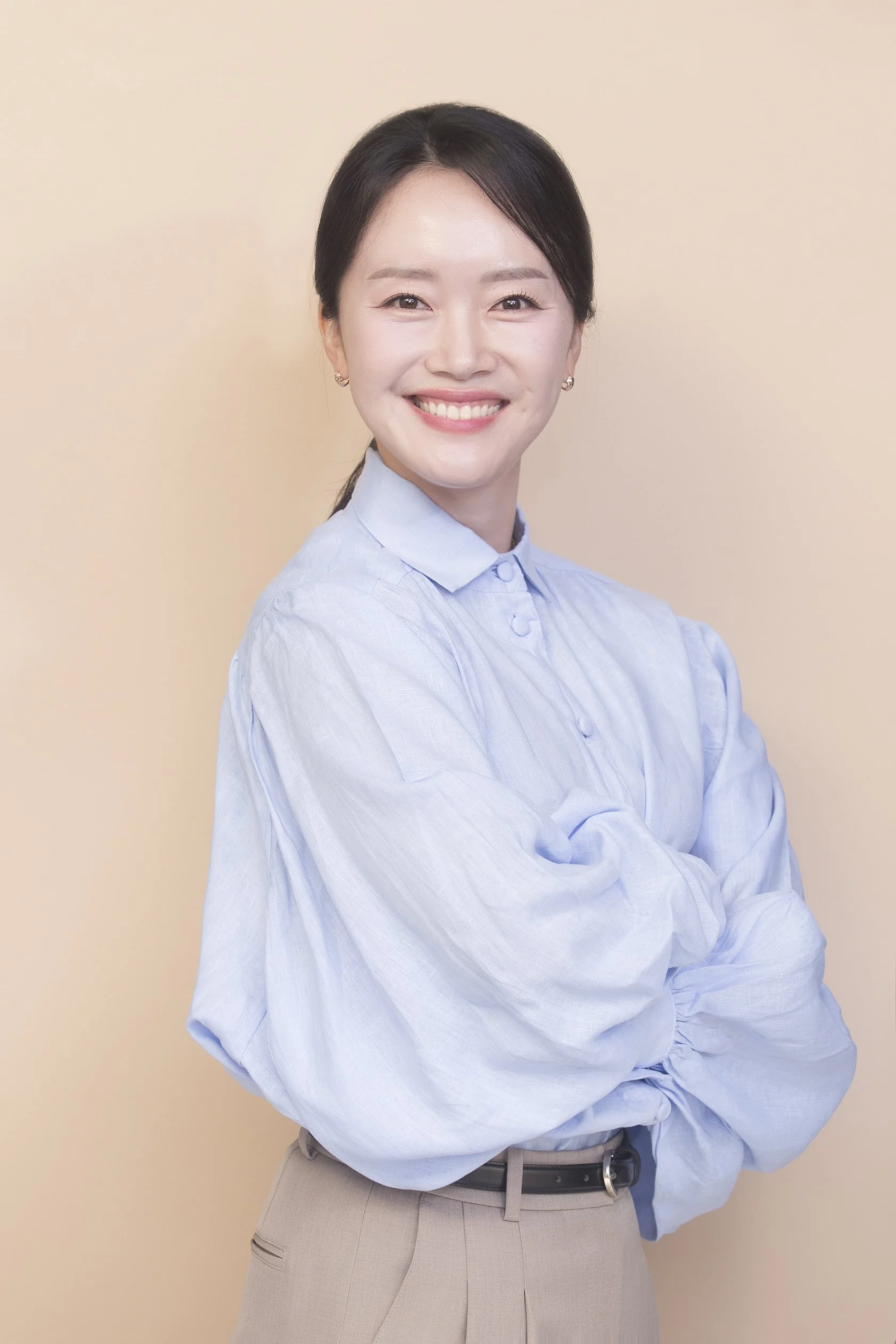 A woman with dark hair smiling, wearing a light blue button-up shirt and beige pants, standing against a plain beige background. She is a CEO, CFO, accountant, chartered accountant (CA ANZ), and MBA graduate.