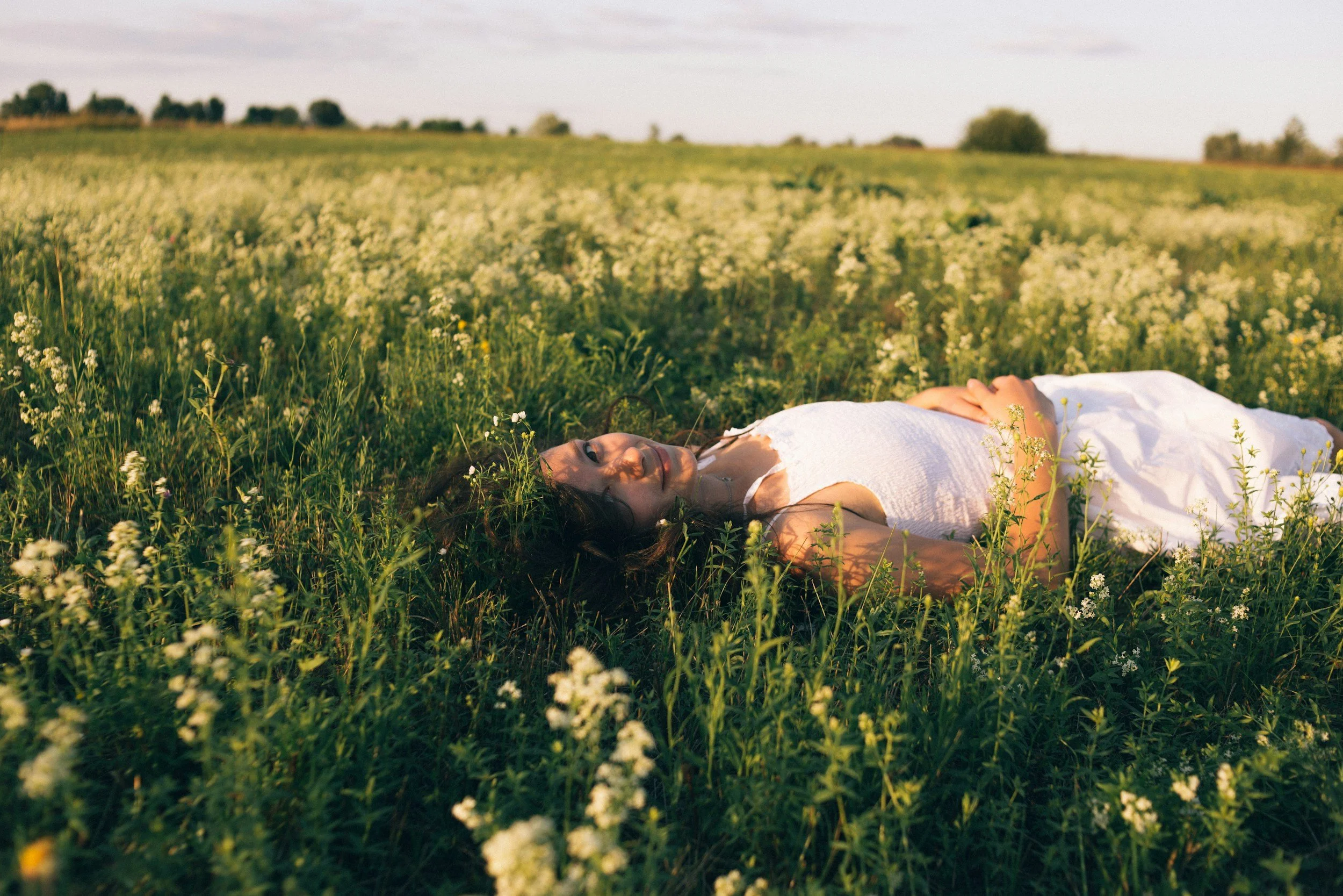 Eine junge Frau liegt entspannt auf einer Wiese mit kleinen weißen Blumen, umgeben von einer weiten Landschaft mit Bäumen im Hintergrund, im Sonnenschein.