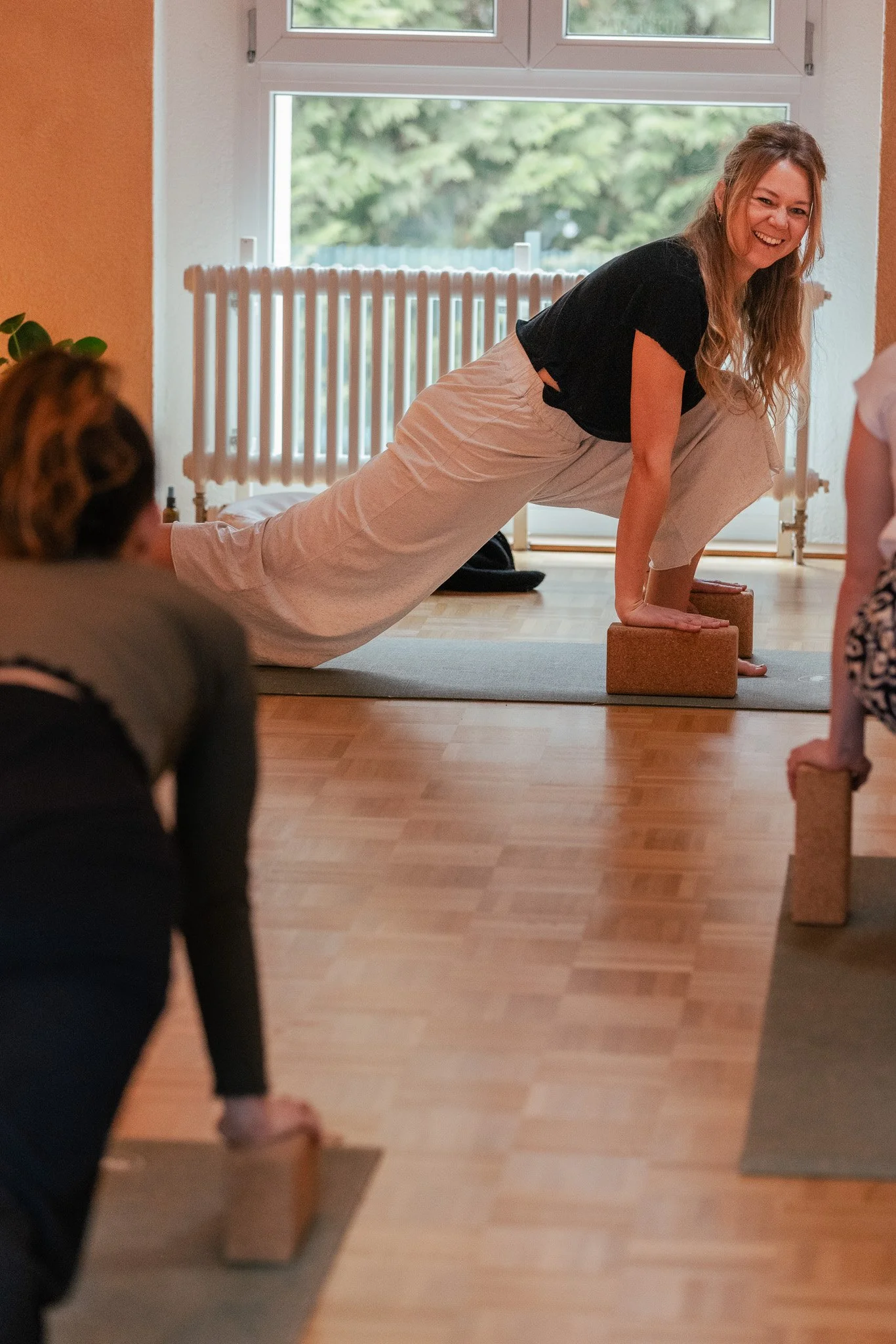 Frau macht Yoga in einem Raum mit großen Fenstern, unterstützt auf zwei Yoga-Blöcken, lächelnd, während andere Teilnehmer mit Yoga-Blöcken auf einer Matte liegen. Traumasensitives Yoga. Somatic Yoga.